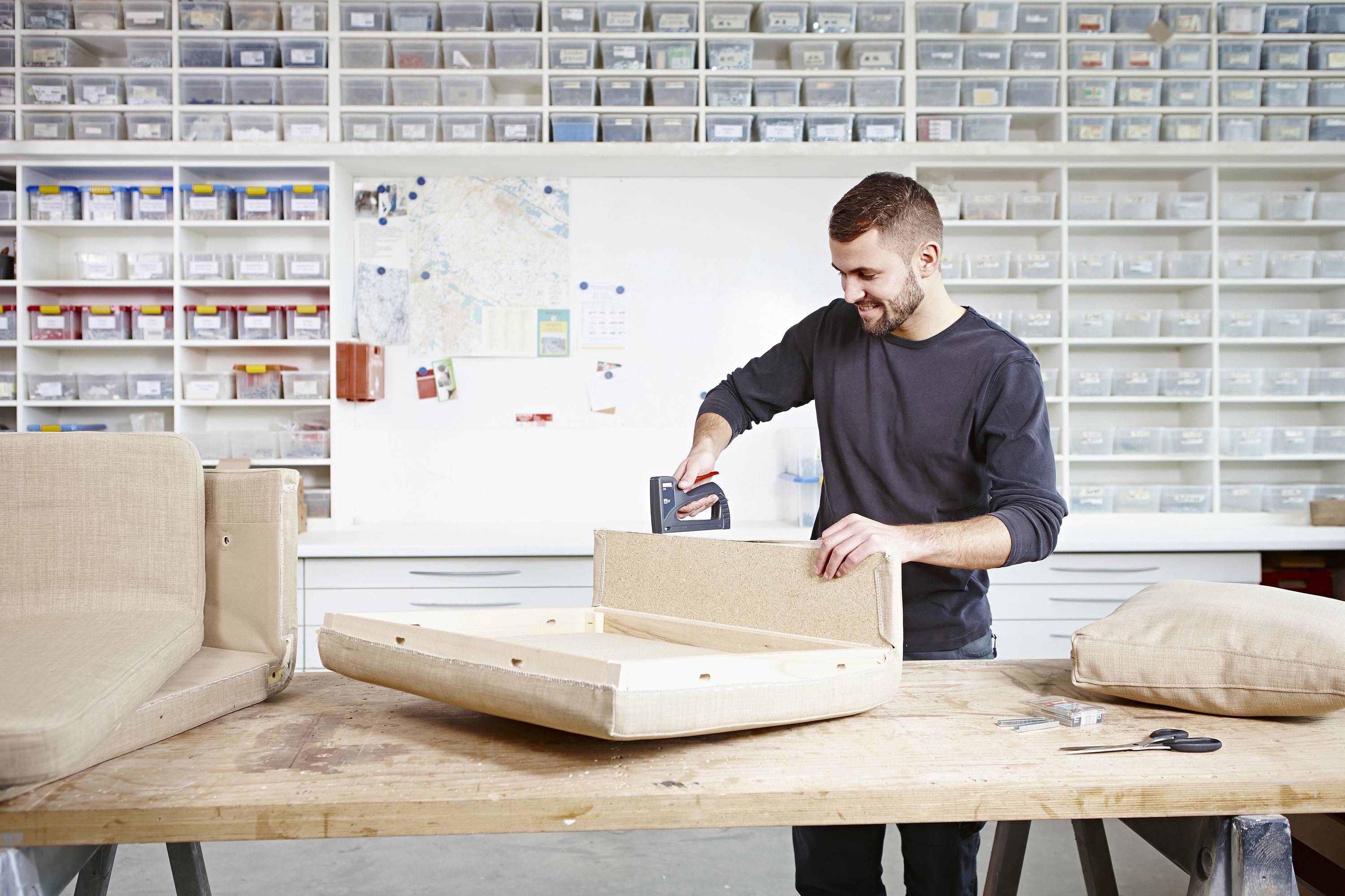 A person in a workshop is upholstering a chair with a staple gun. Shelves with boxes of various sizes are in the background.