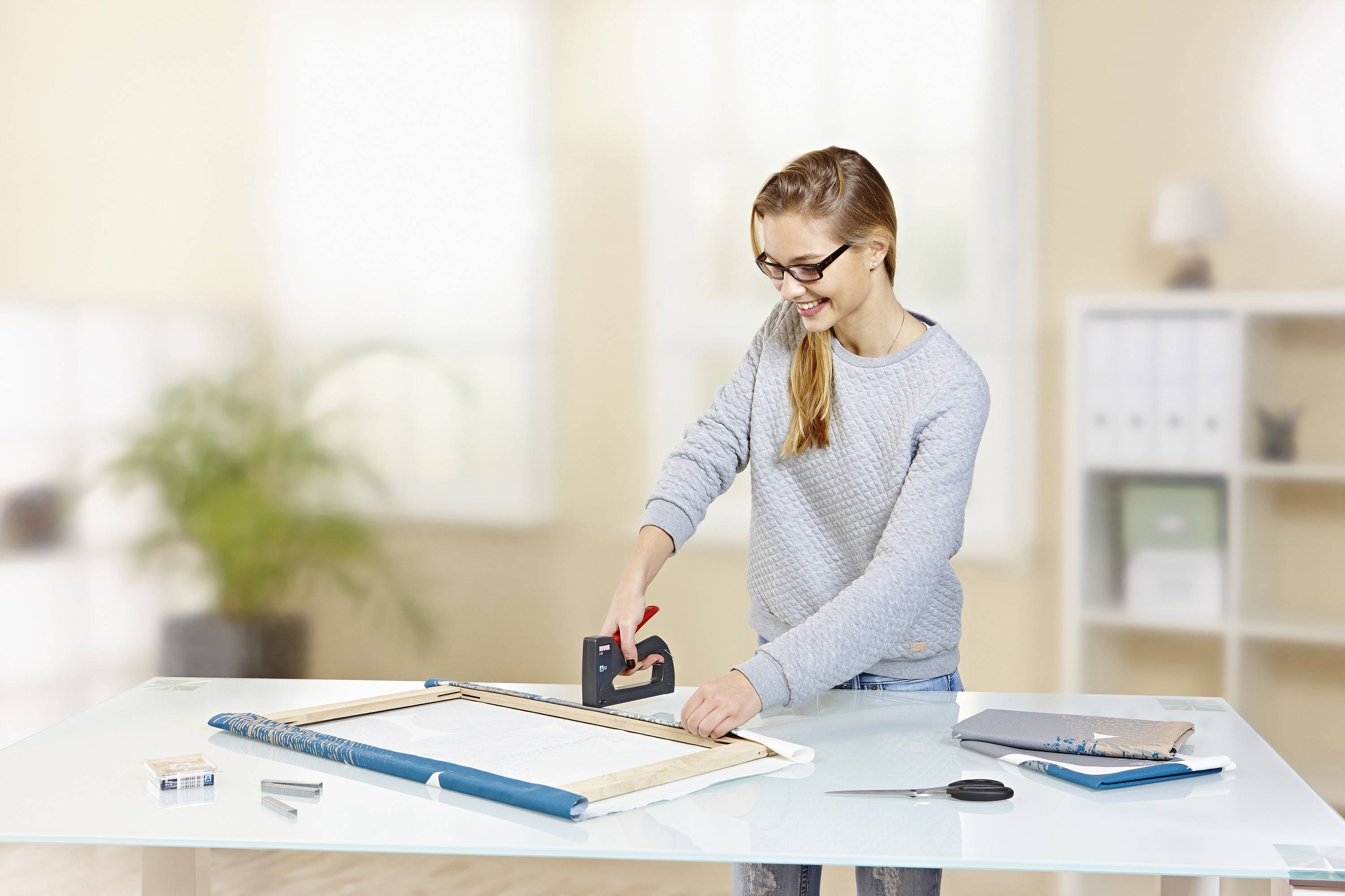 A woman wearing glasses is attaching fabric to a wooden frame using a staple gun in a bright office with a desk.