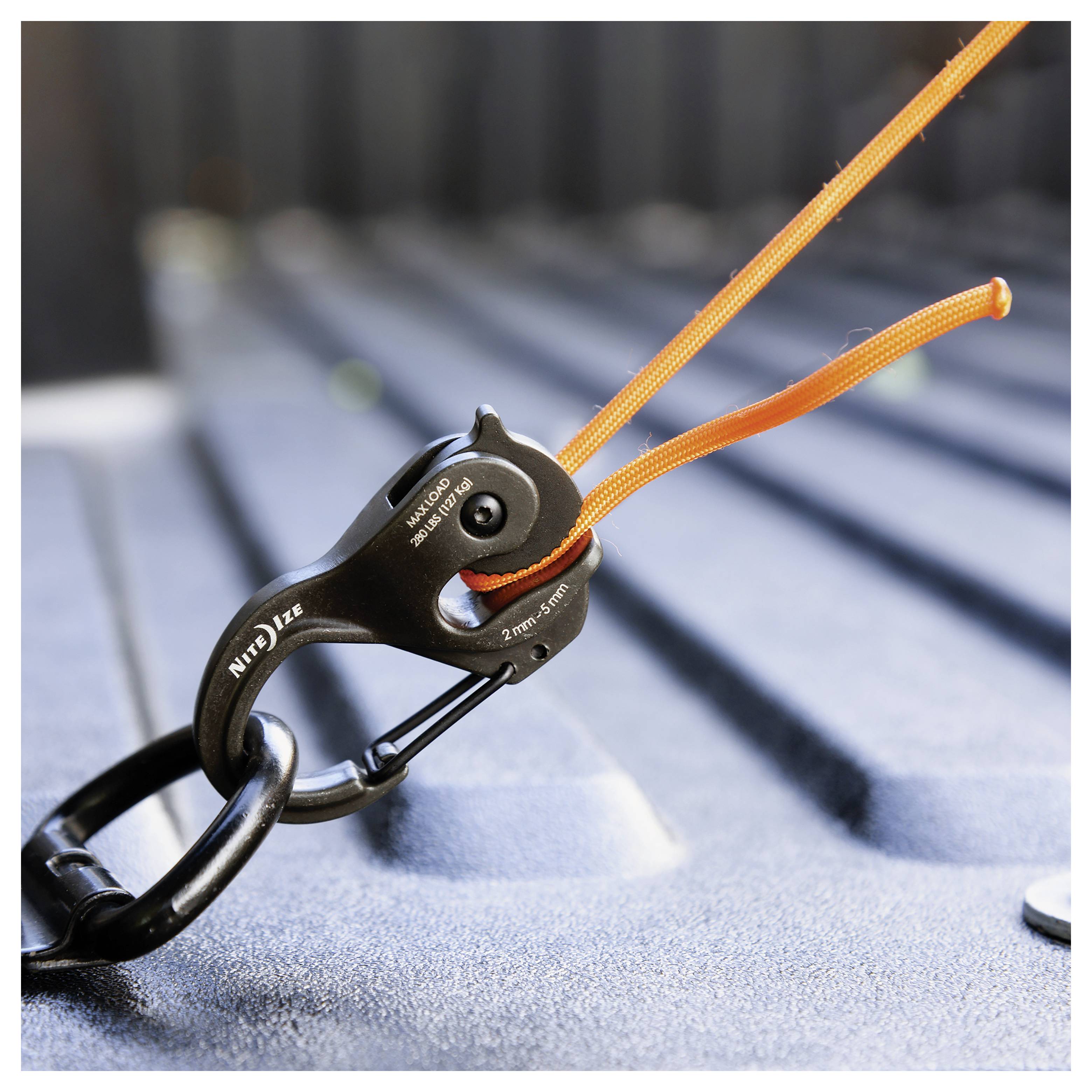 A black metal pulley with an orange rope looped through it, secured to a textured gray surface. The setting suggests tensioning or load adjustment.