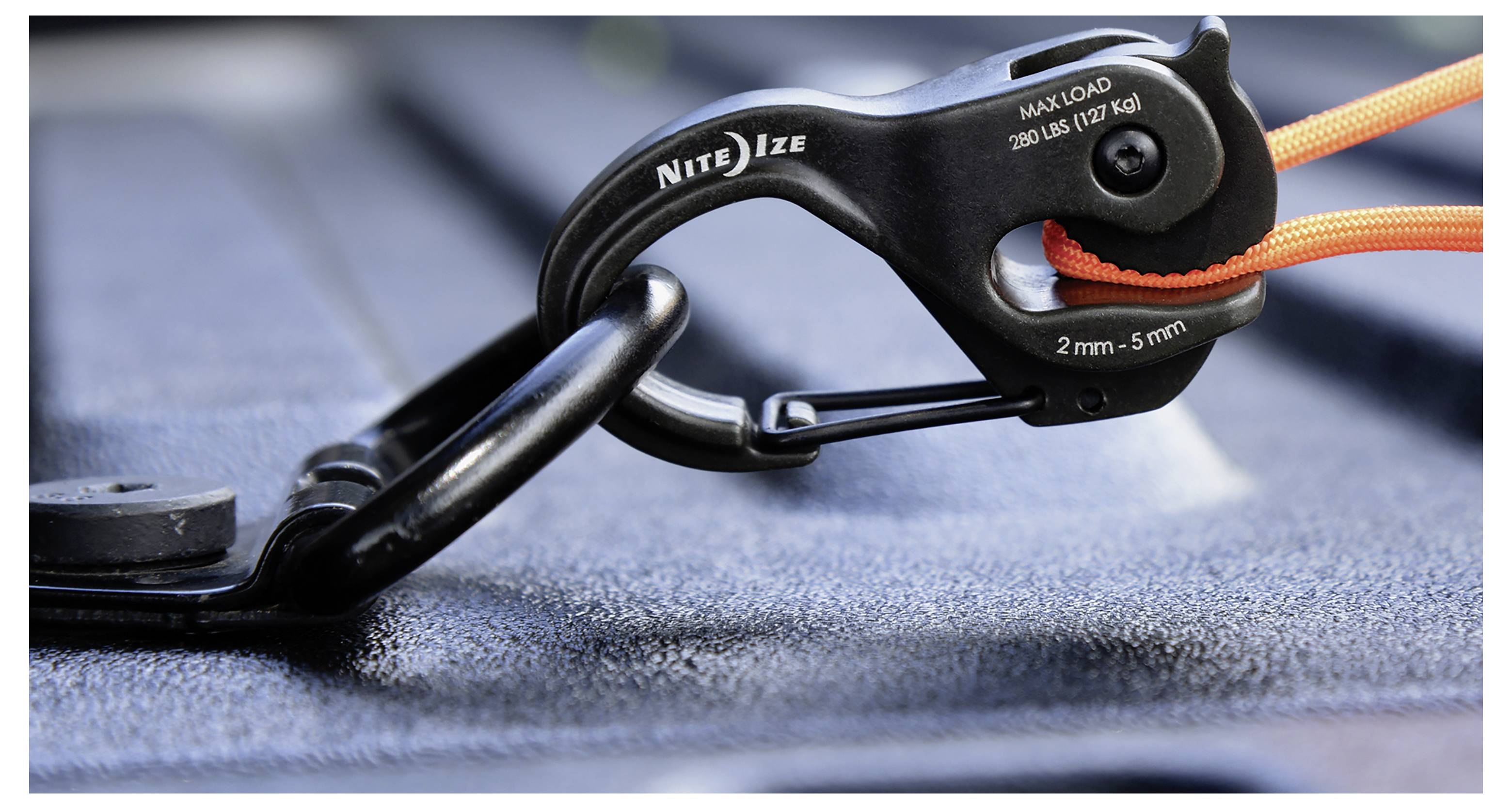 A close-up of a black Nite Ize clip securing an orange rope to a metal anchor point. The clip is labeled 'Max Load: 50 lbs (22 kg)'.