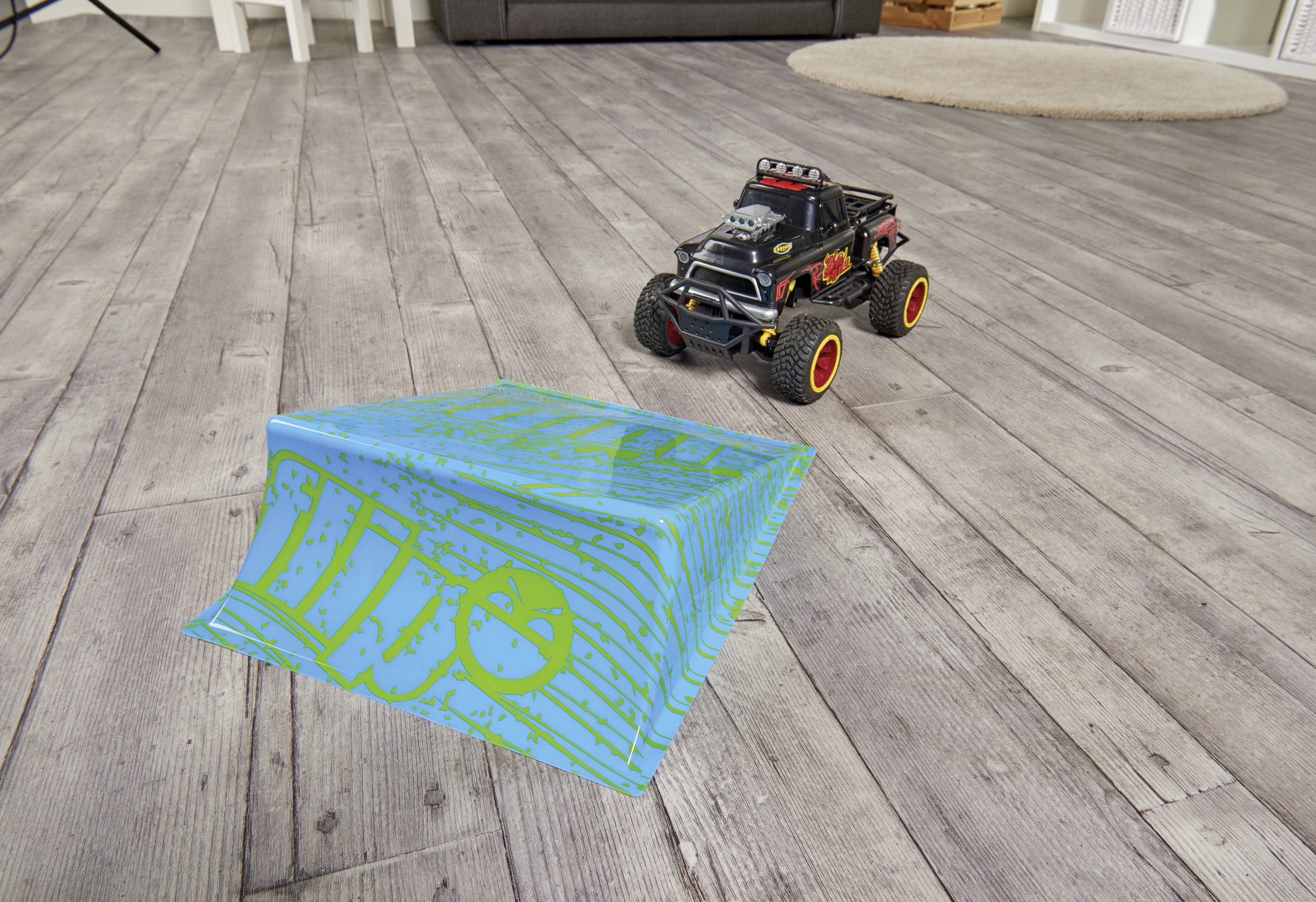 A black toy car with red tyres drives over a blue-green ramp in a living room with wooden flooring.
