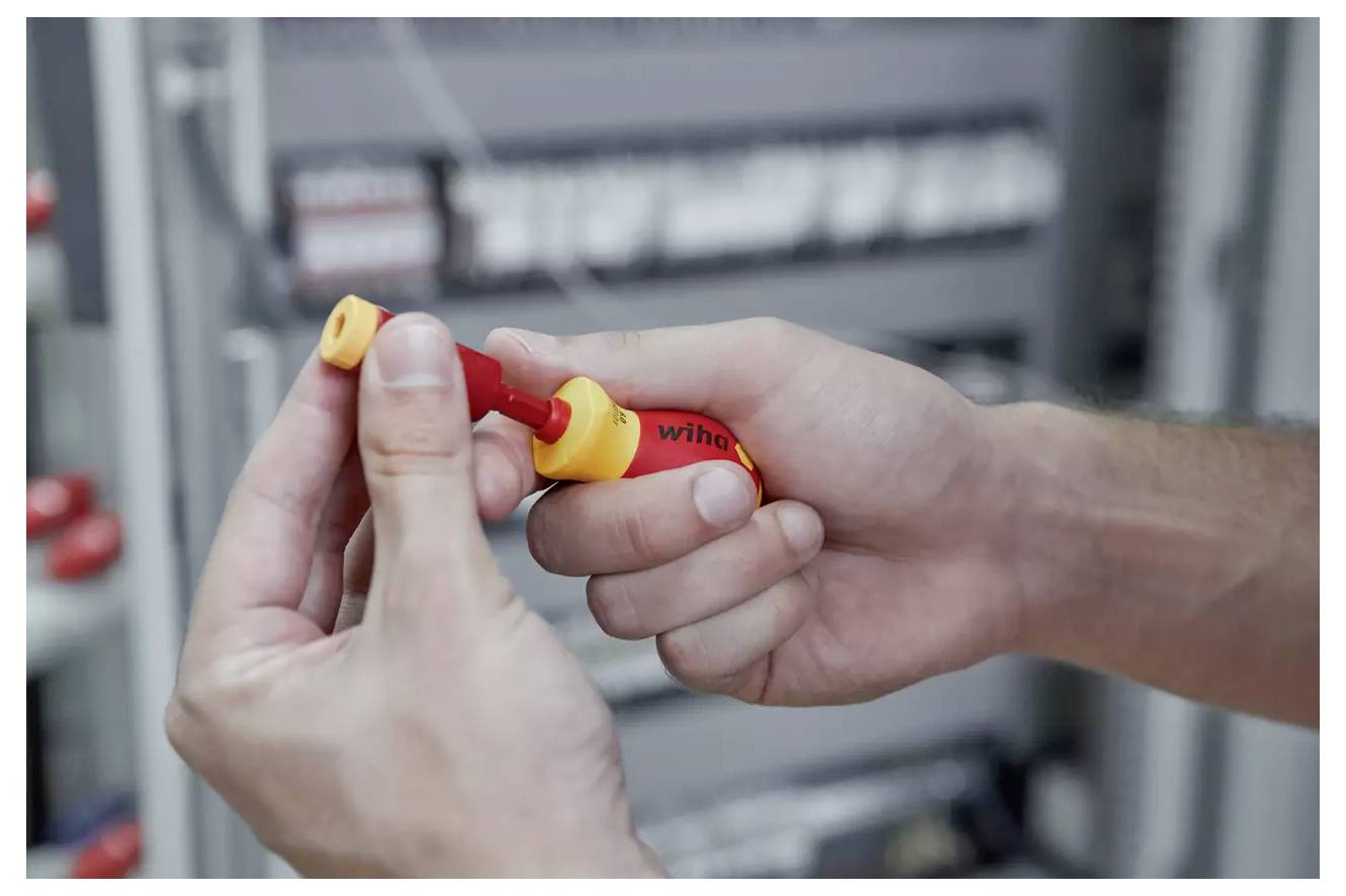A person is holding a red and yellow electrical testing screwdriver in front of a blurred electrical panel background.