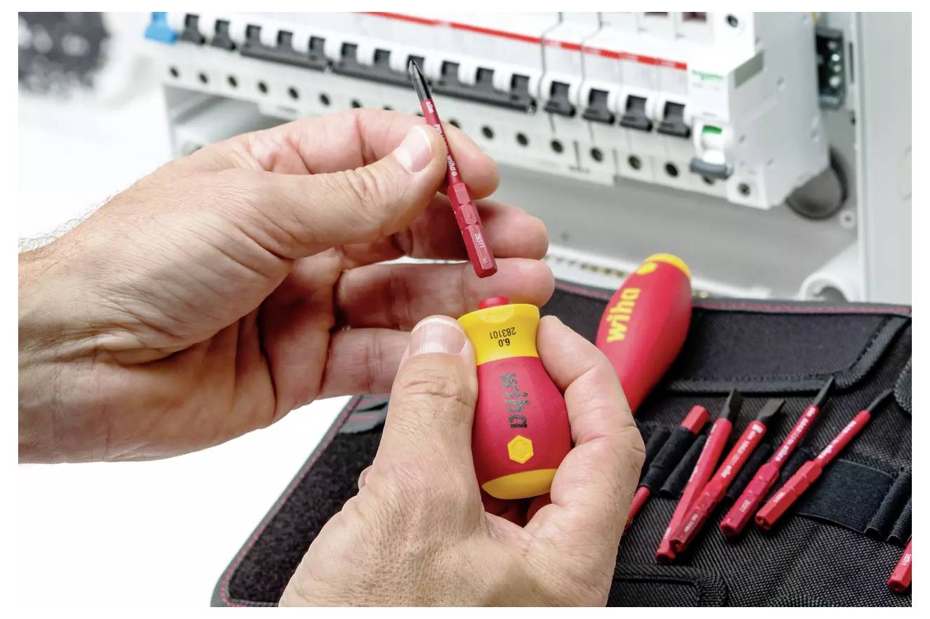 A pair of hands holding a red and yellow screwdriver set in front of an electrical panel, with additional screwdriver bits visible nearby.