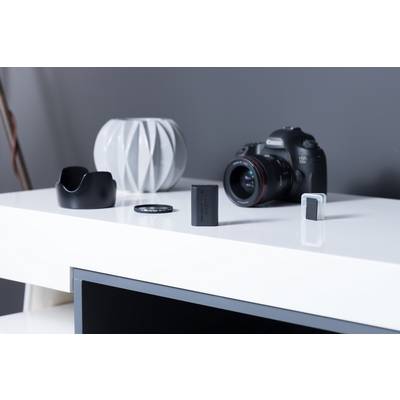 A camera with a lens sits on a white table. Beside it are a lens cap, a battery pack, and a grey decorative item.