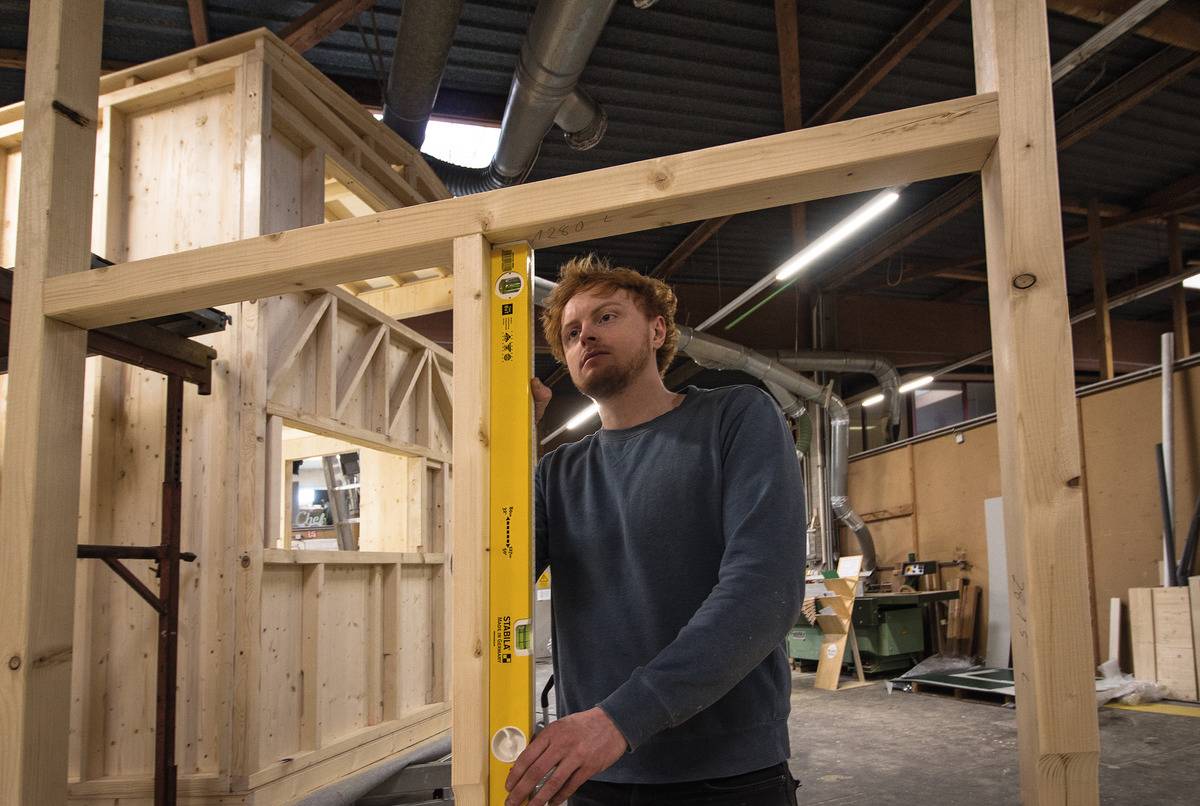 A young man is using a spirit level to align a wooden frame precisely in a workshop. Wooden structures are visible in the background.