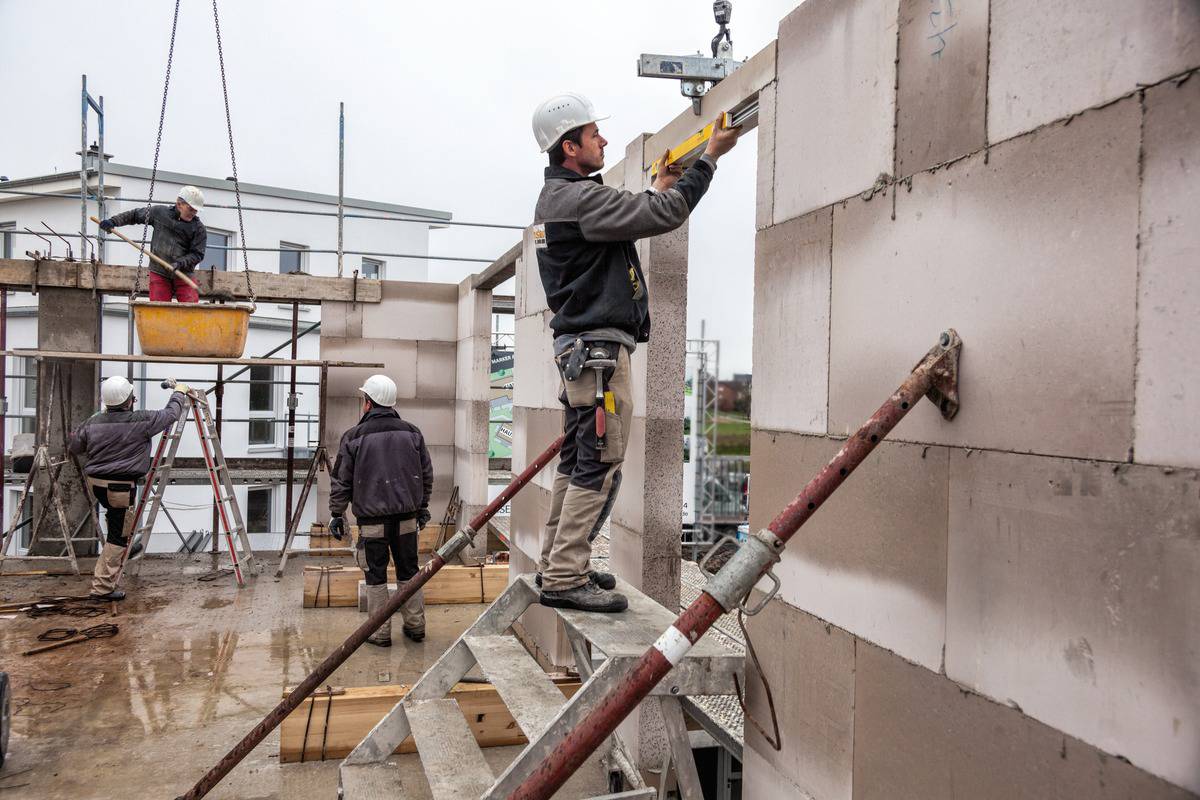 Construction workers on a building site, wearing hard hats and working on a wall. One man on a ladder is checking the alignment with a spirit level.