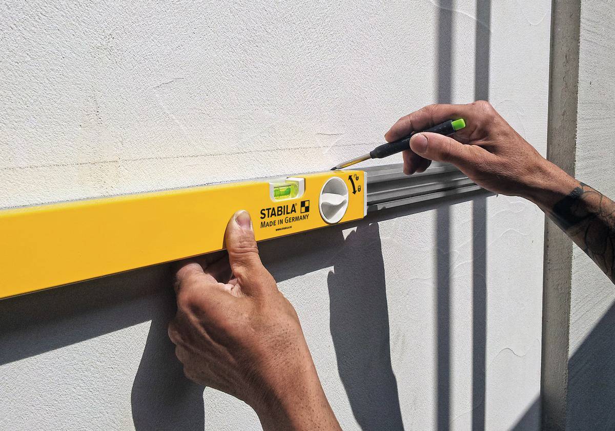 A person is using a spirit level on a white wall to mark a straight line. The spirit level is yellow and made in Germany.