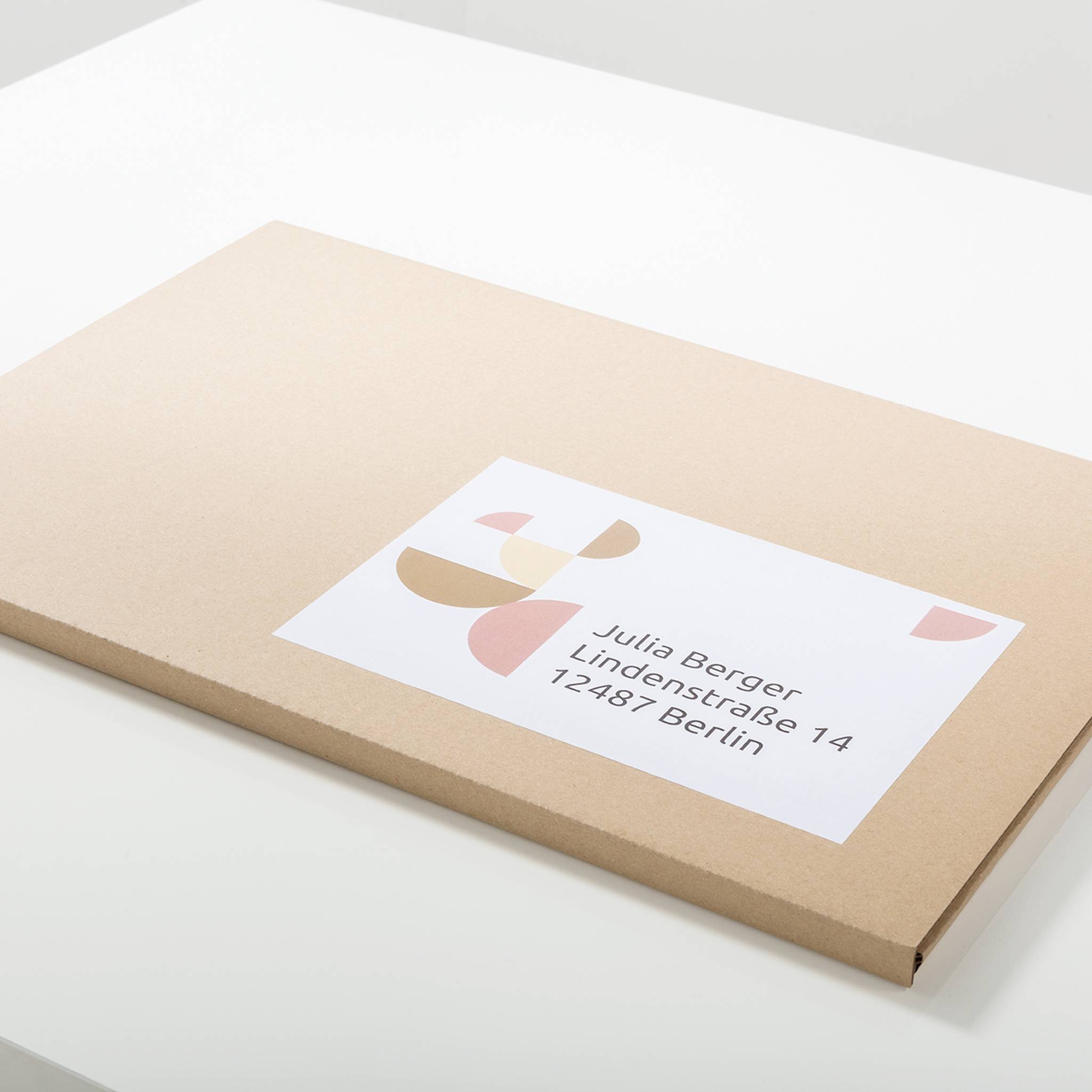 A flat, beige envelope is lying on a table. It has a label with the name 'Julia Berger' and the address 'Lindenstraße 14, 12487 Berlin'.