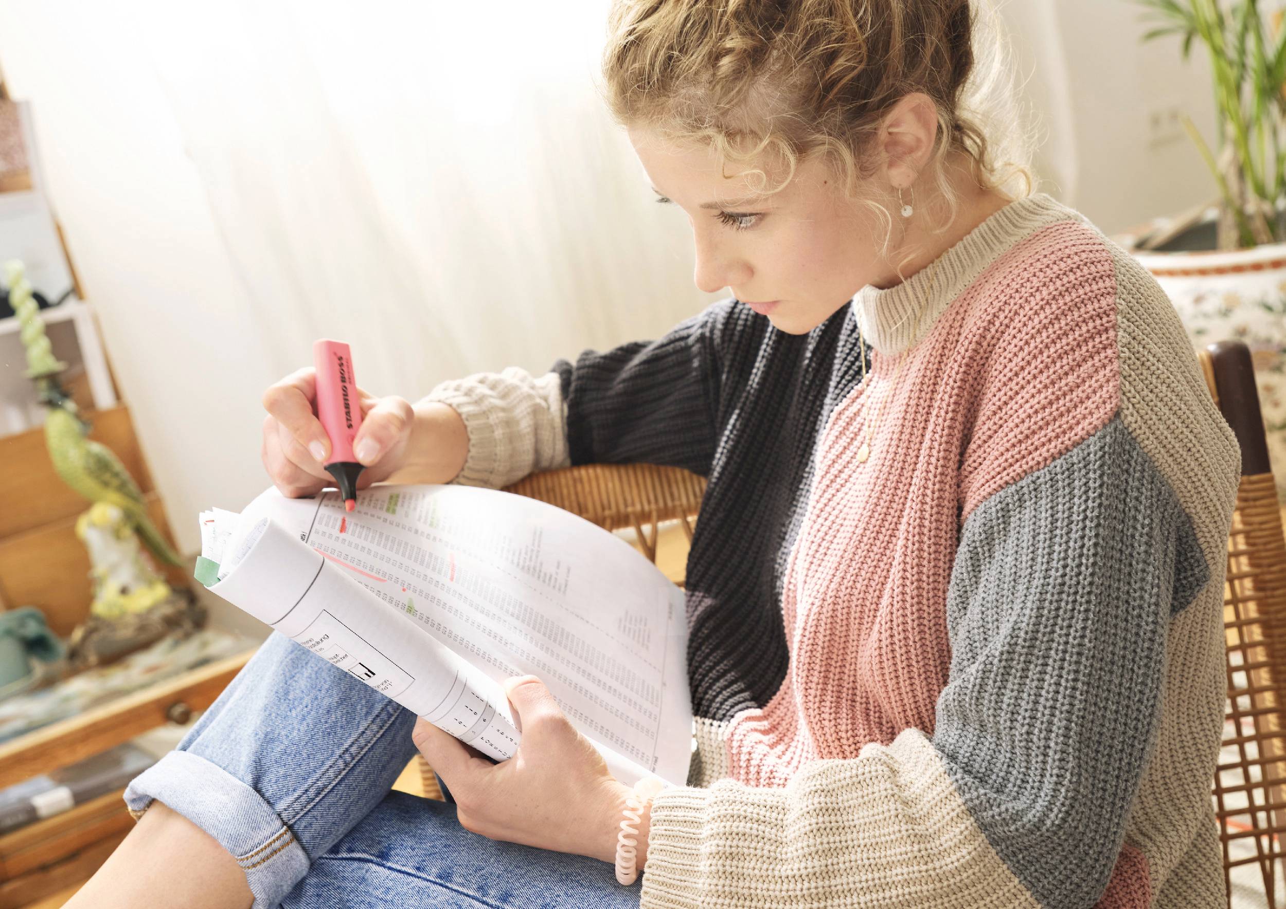 A woman is sitting in a chair and highlighting passages with a highlighter in a document. She is wearing a colourful knitted jumper.