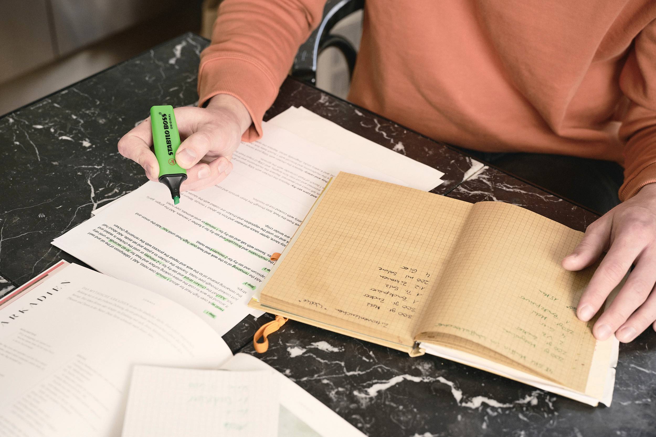 A person in an orange jumper is highlighting text on a printout. An open notebook lies beside it.