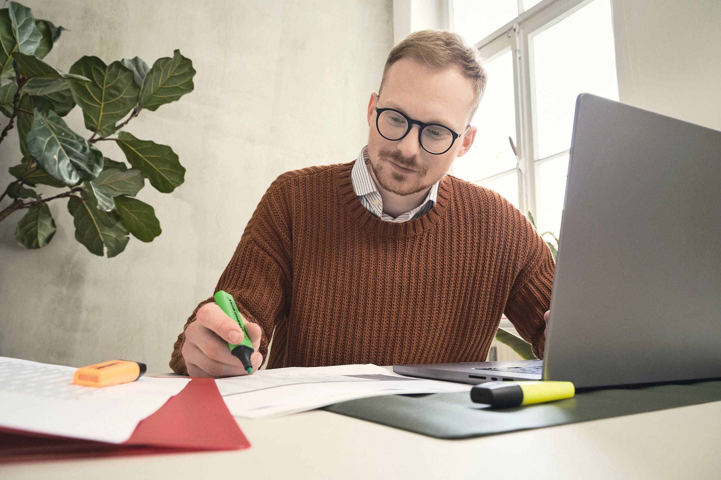 A man wearing glasses sits at a desk, working intently with a laptop and documents. A plant is positioned in the background.