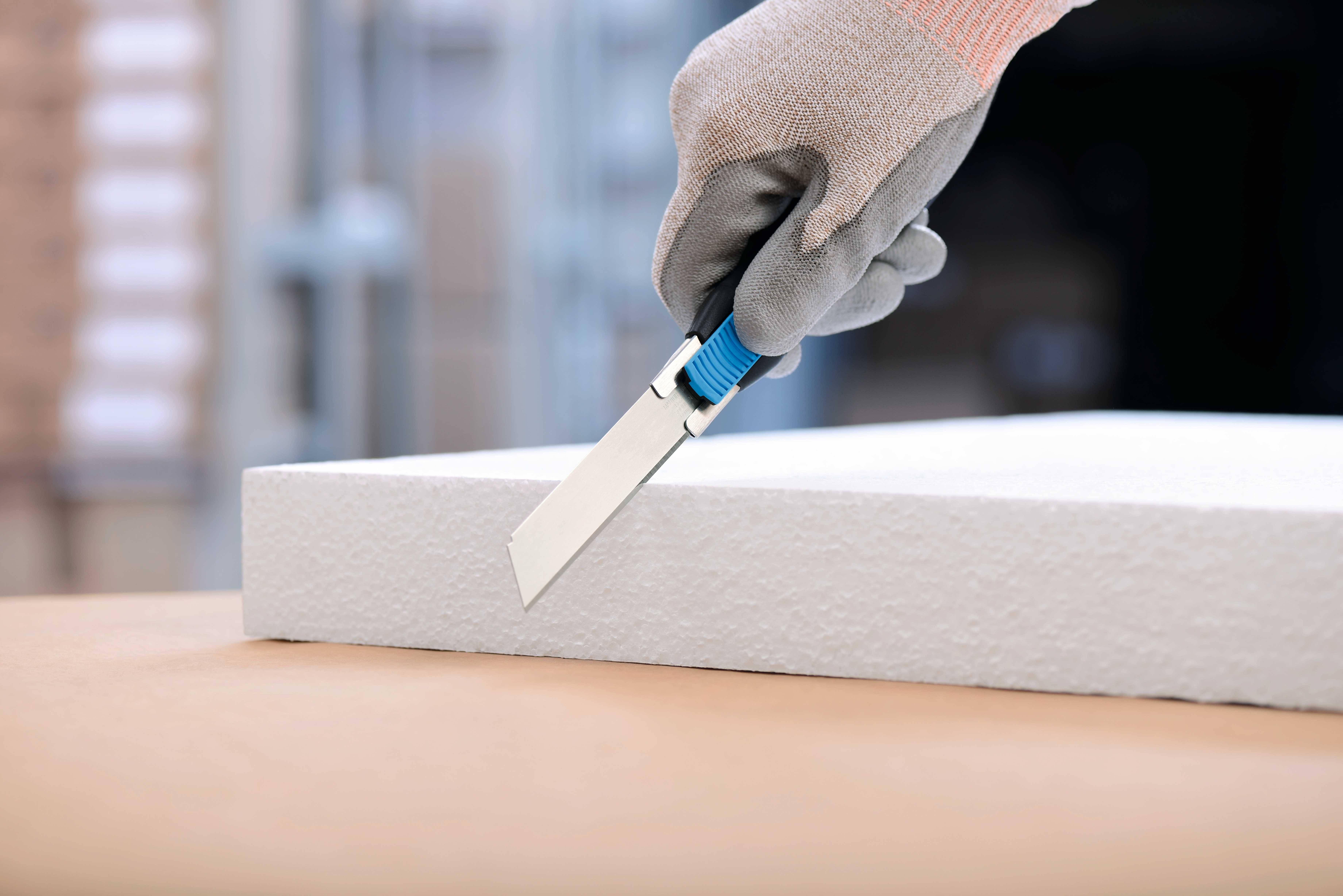 'A hand wearing a grey glove cuts a white polystyrene sheet with a craft knife on a table.'