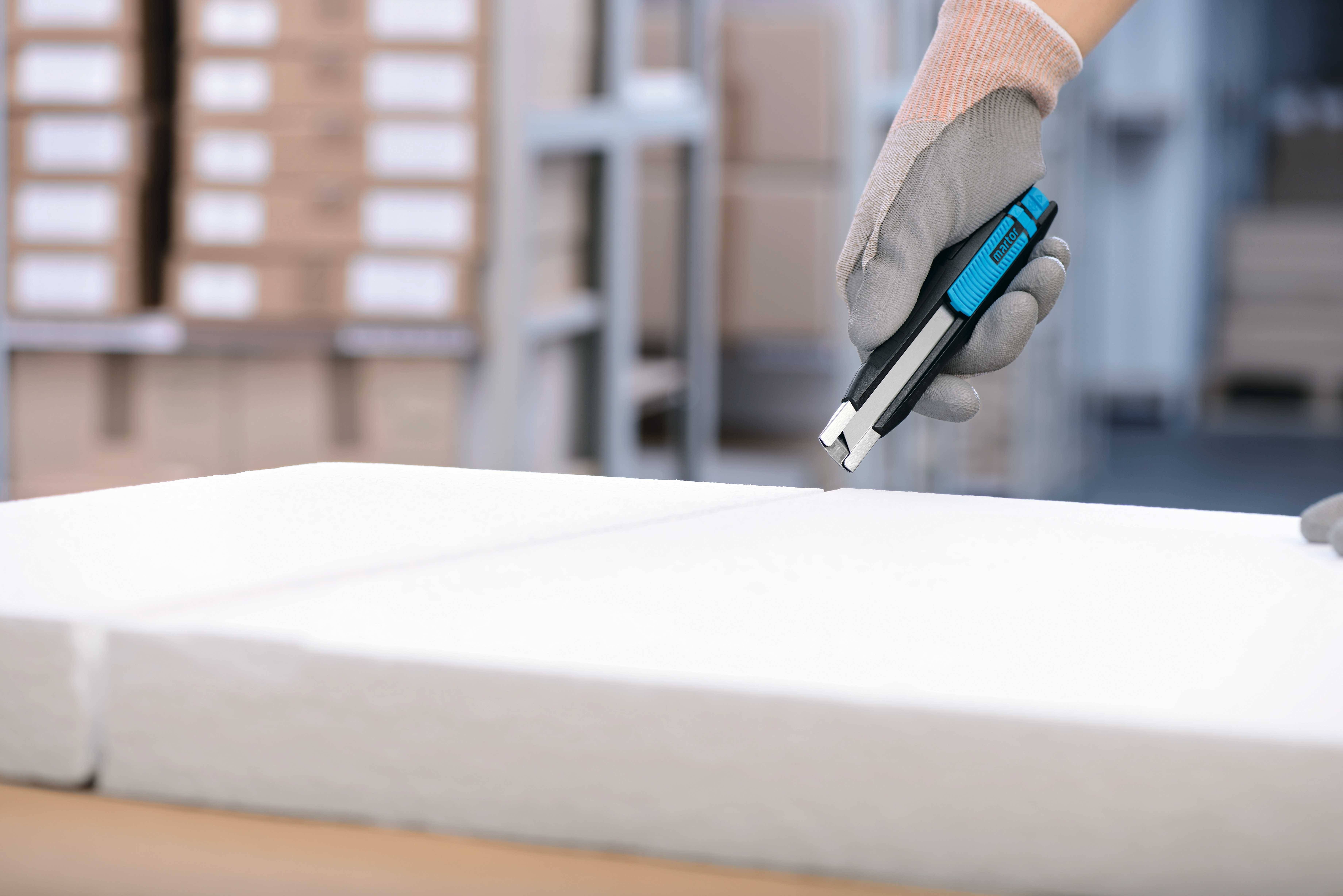 A gloved hand holds a carpet knife and is cutting polystyrene in a warehouse setting. Shelves can be seen in the background.