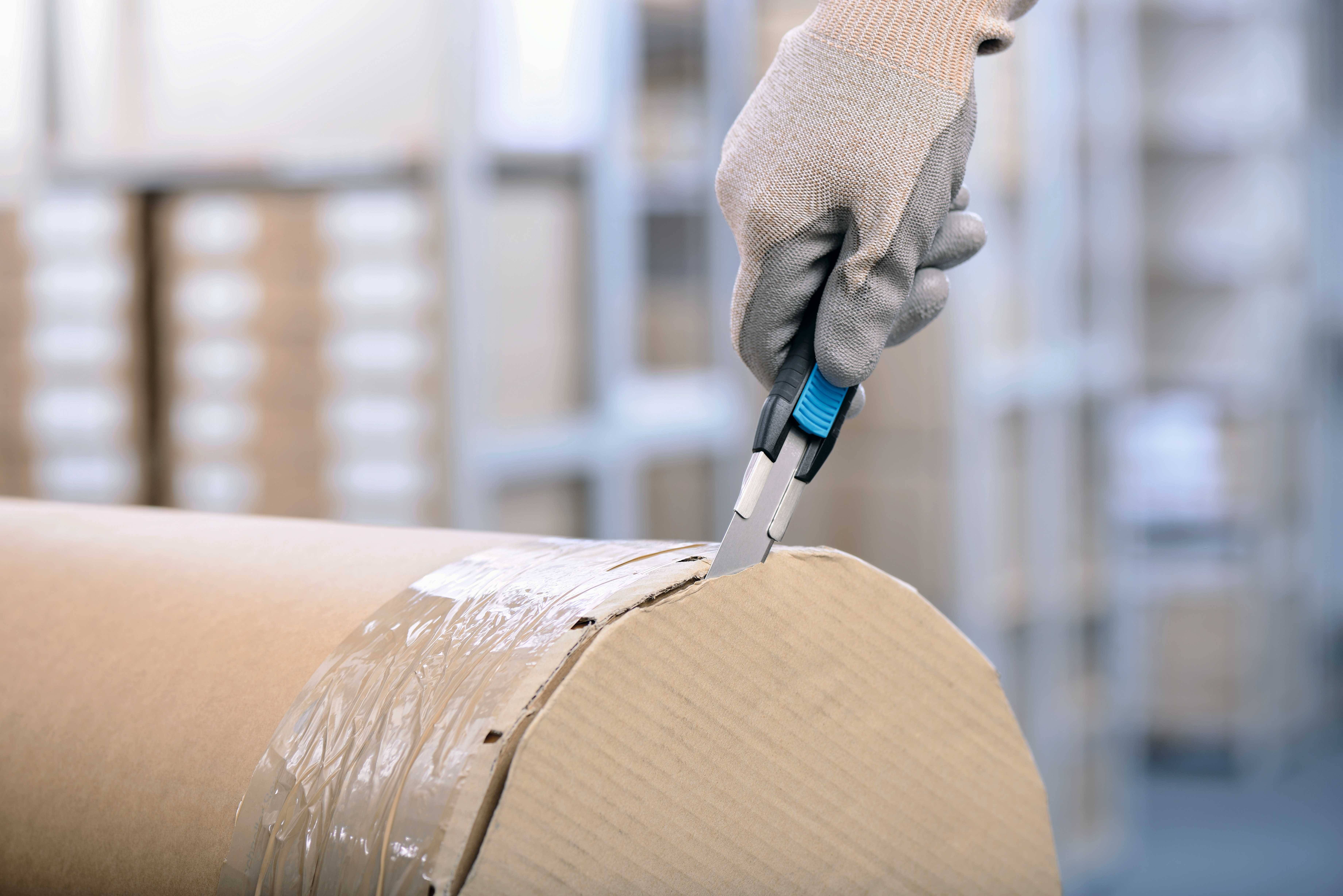 A person is cutting open a cardboard-packaged roll using a utility knife. Background: blurry shelves with packages.