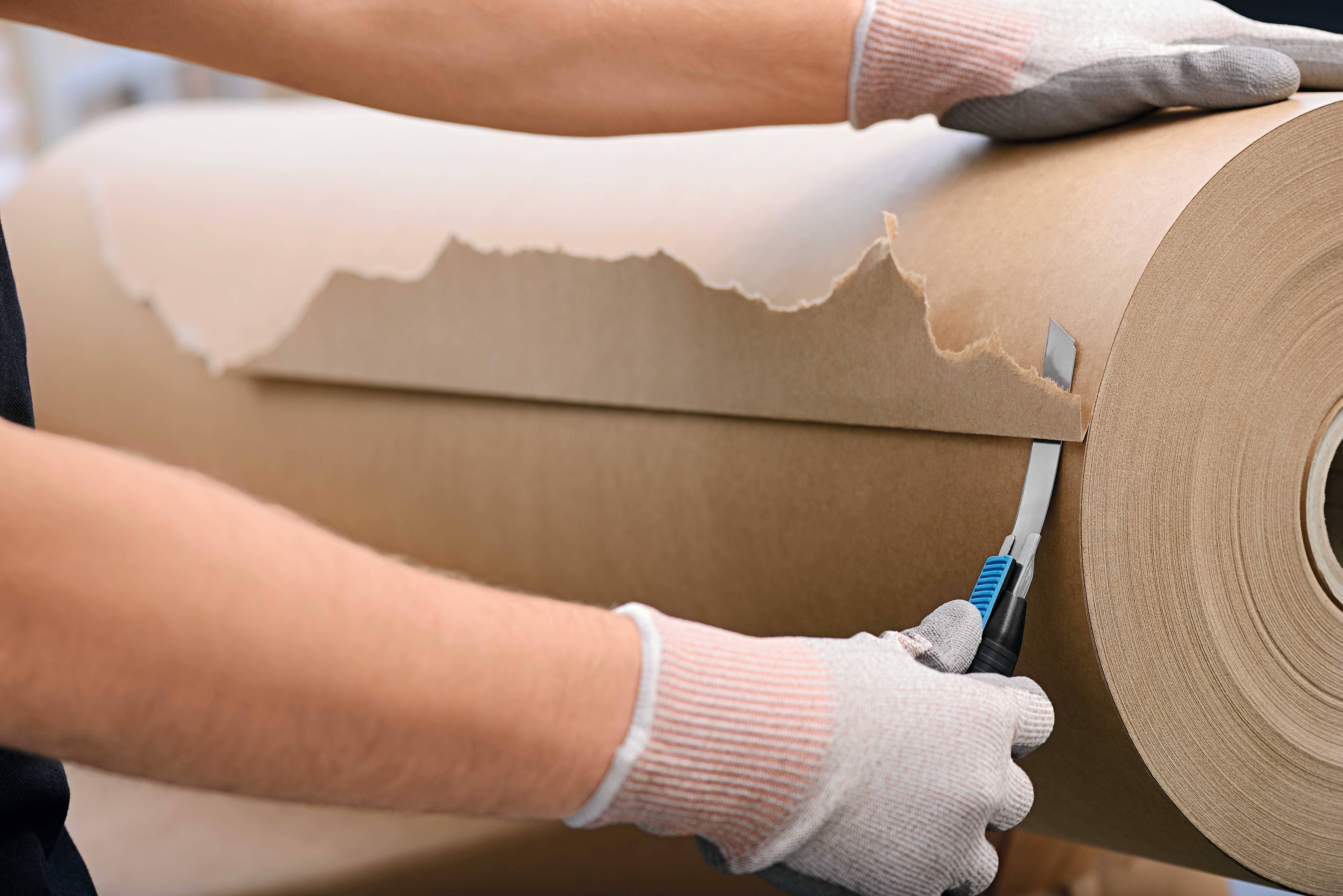 A person wearing gloves is cutting a roll of thick paper with a knife.
