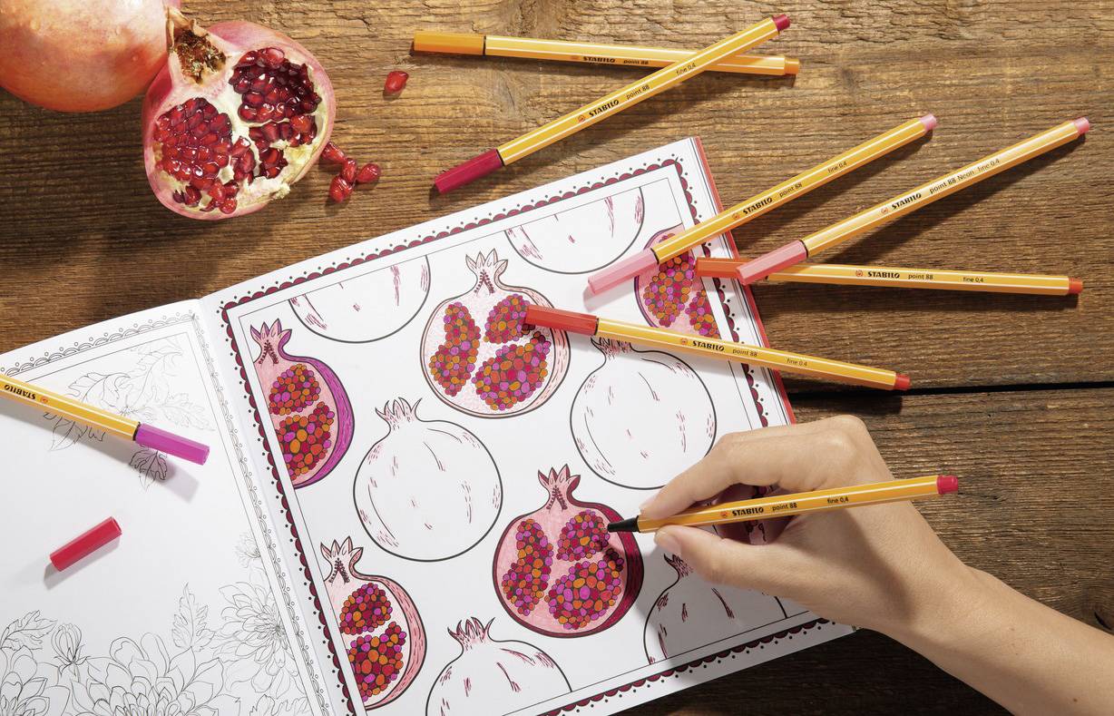 A colouring book shows drawn pomegranates. A hand is colouring a pomegranate with pink and red pencils. A real pomegranate lies on top.