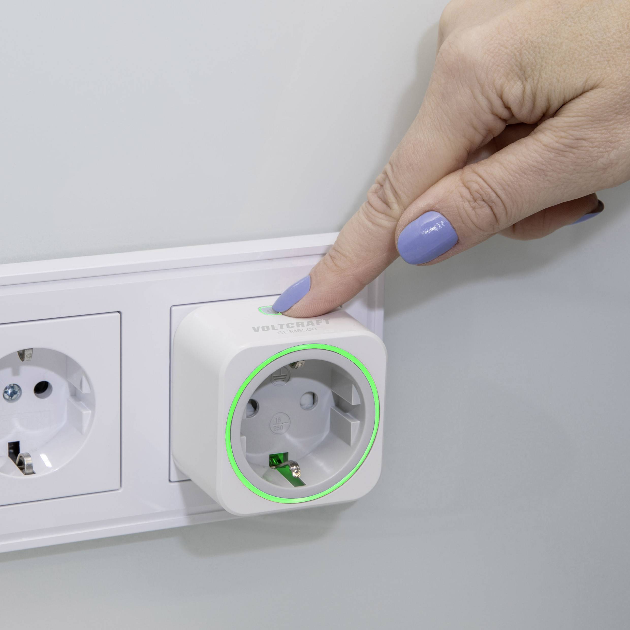 A hand presses a smart plug that is plugged into a socket. The plug glows green, indicating it is ready for use.