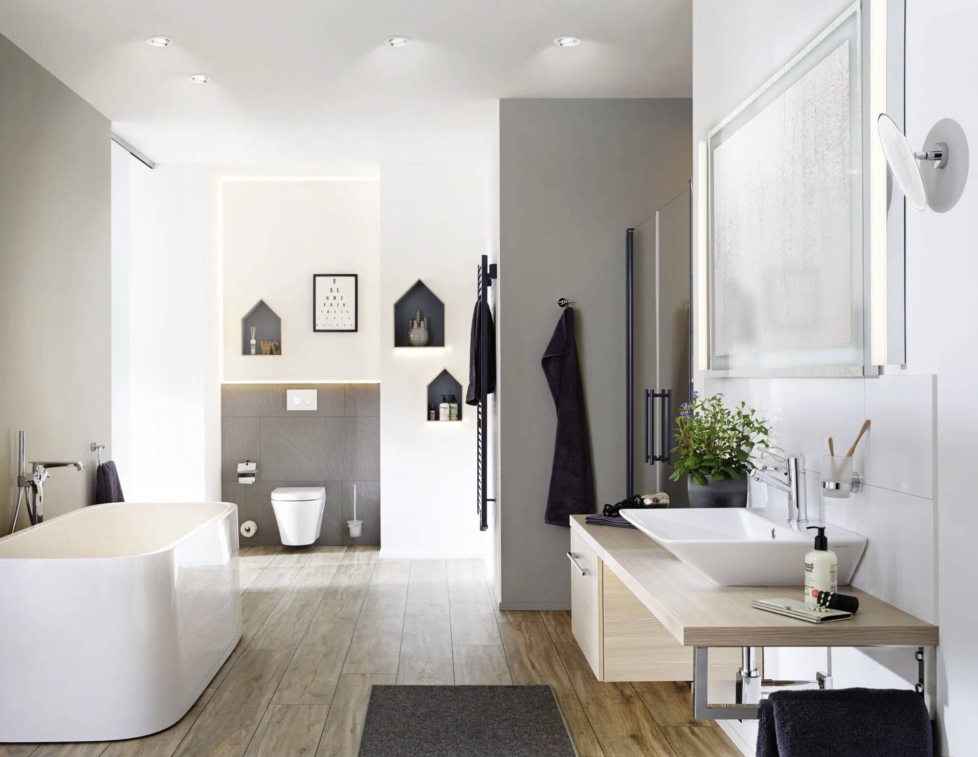 Modern bathroom with a freestanding tub, wall-mounted toilet, two sinks on a wooden counter, and decorative shelves with framed art.