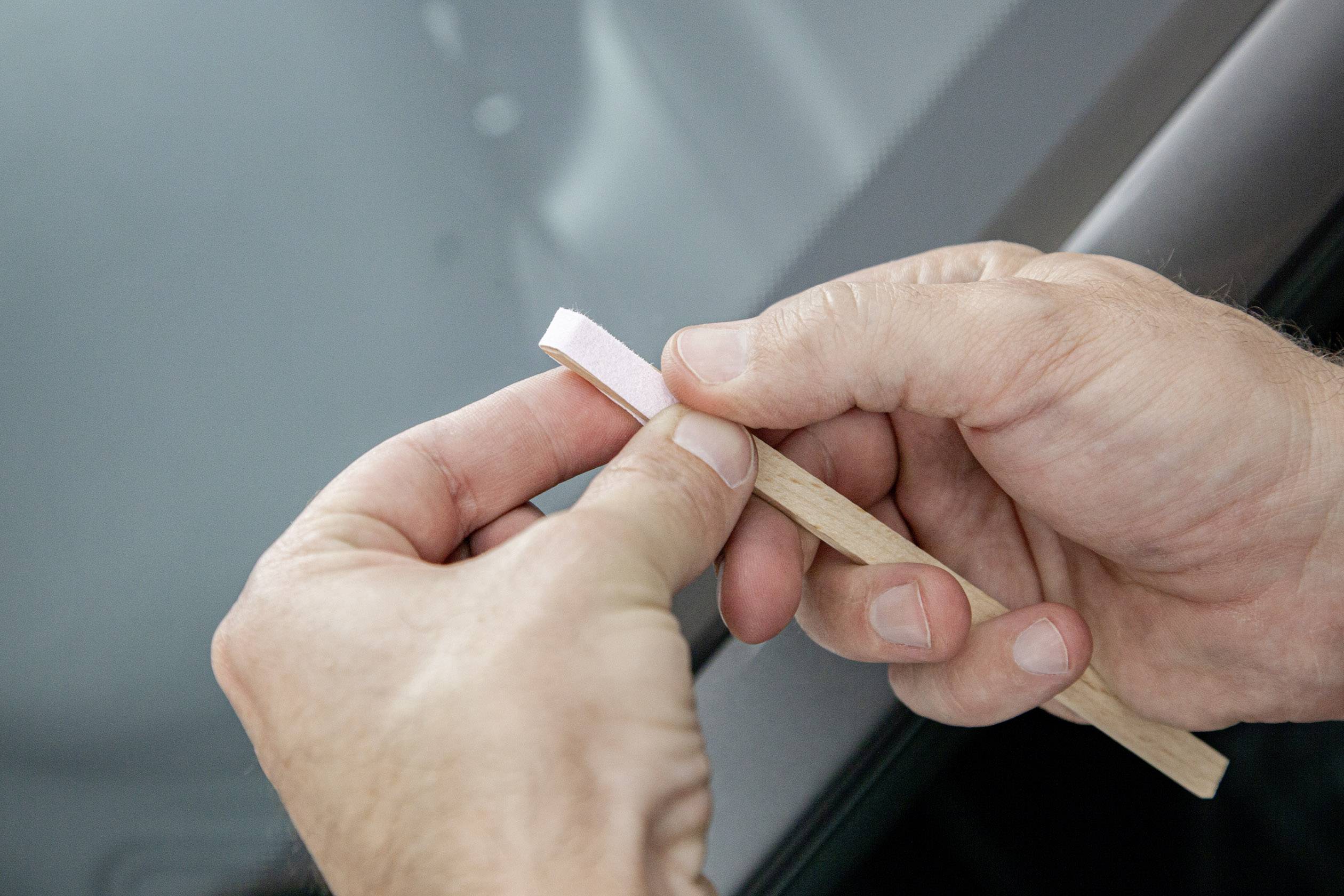 A hand is holding a small wooden stick with a thin adhesive strip. The other hand is peeling off the protective film. The background is a car window.