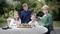 Four smiling children are sitting around a round table outdoors, with a chessboard on the table and trees in the background.