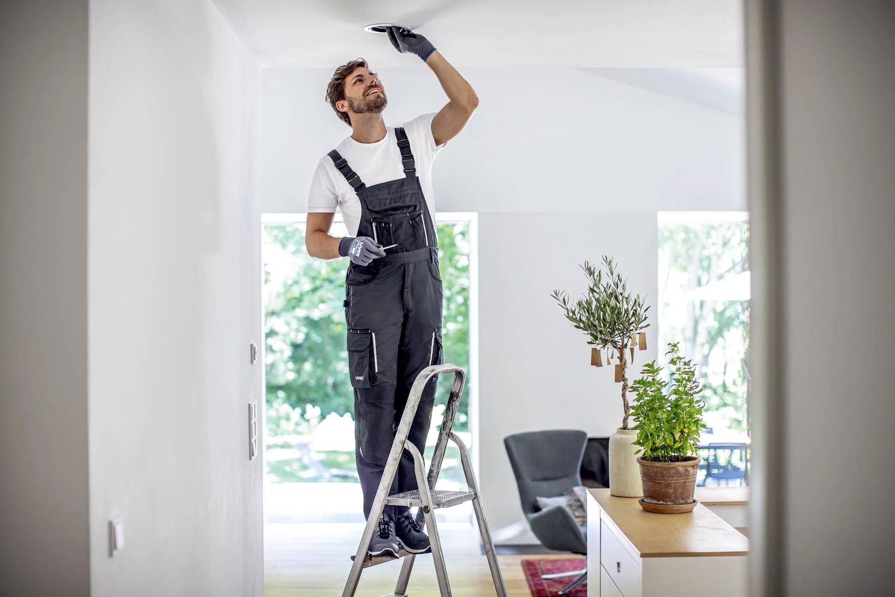 A man in dungarees is standing on a ladder and repairing the ceiling in a bright room.
