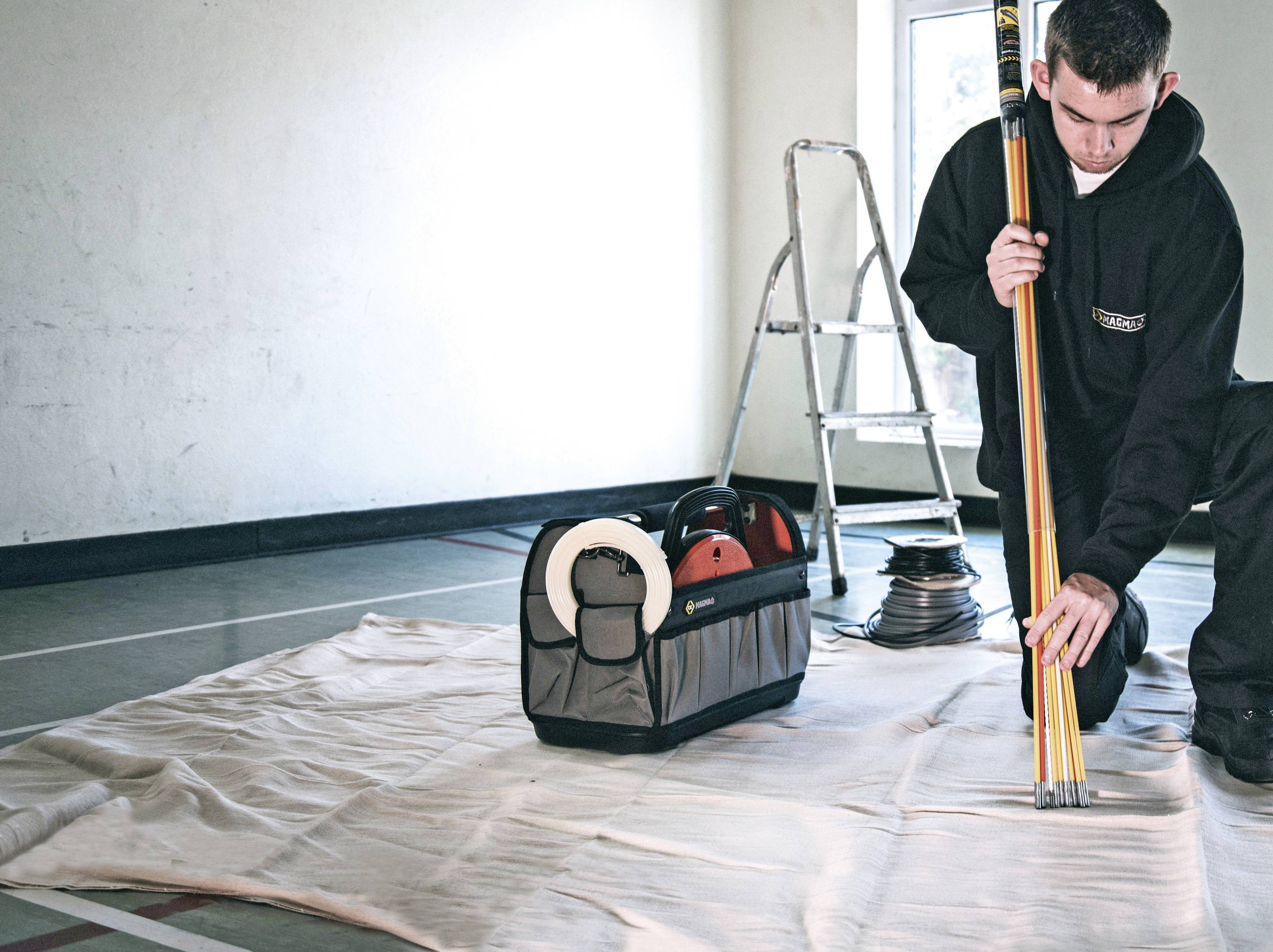 A tradesman is arranging measuring laths in front of a ladder in a room. Next to him, a bag with cables and tools sits on a tarpaulin.