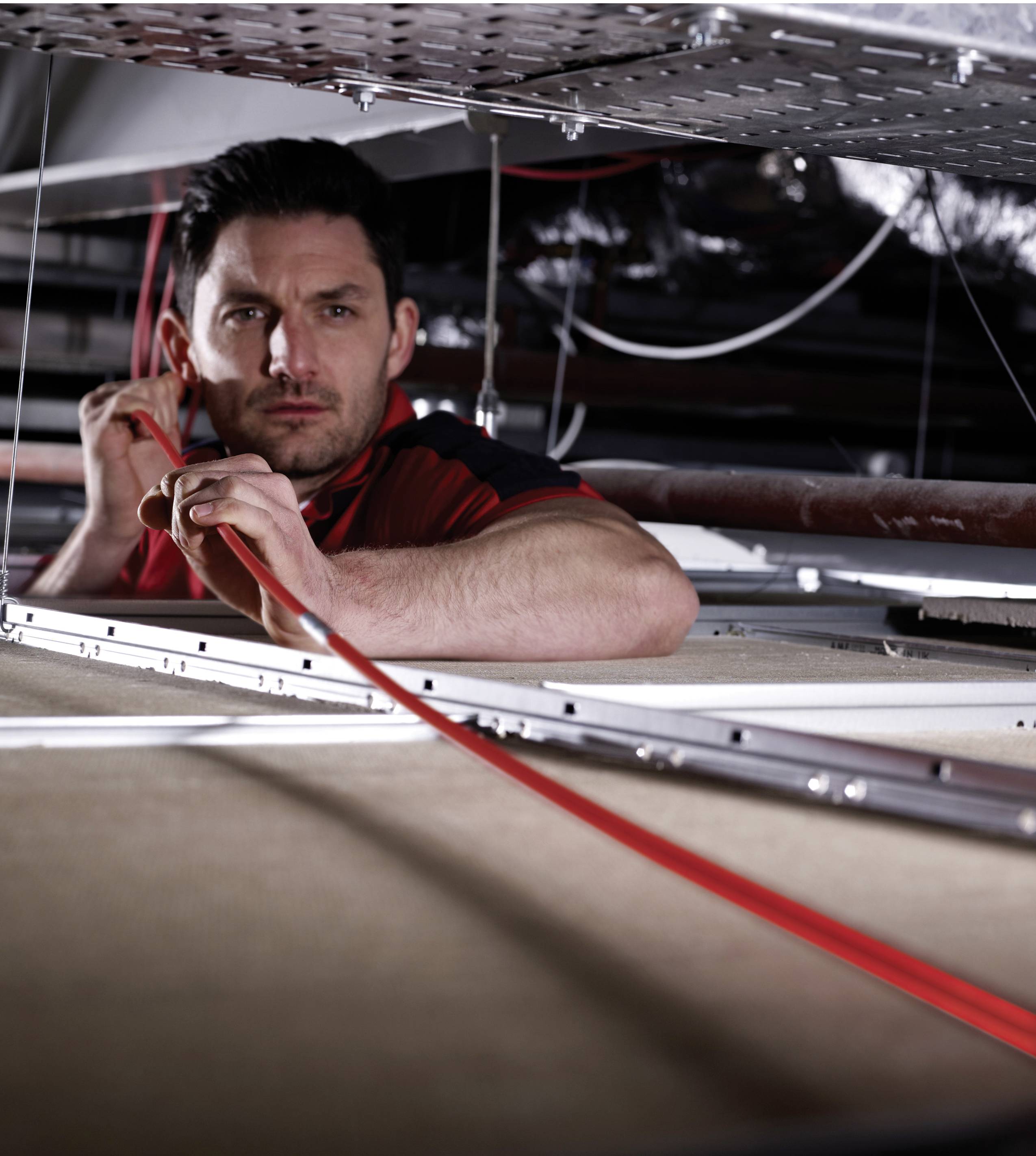 A man is working intently in an industrial setting, threading a red cable through a ceiling.