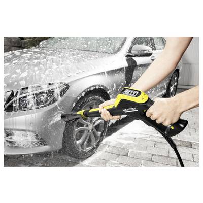 A person's hands using a Kärcher pressure washer, spraying soap and water on a silver car, effectively cleaning it on a driveway.