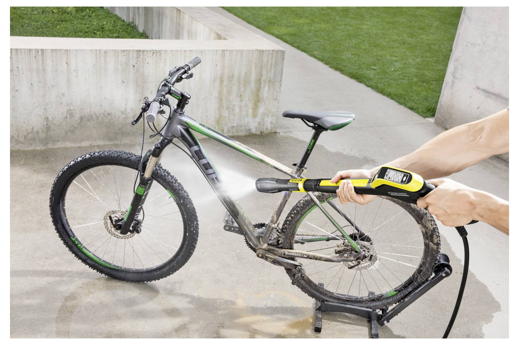 A person uses a high-pressure water hose to clean a muddy bicycle on a concrete surface, with a grassy area in the background.