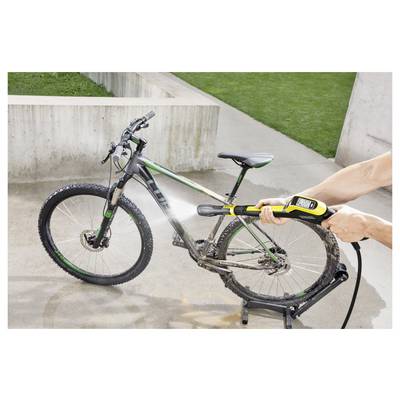 A person uses a high-pressure water hose to clean a muddy bicycle on a concrete surface, with a grassy area in the background.