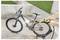 A person uses a high-pressure water hose to clean a muddy bicycle on a concrete surface, with a grassy area in the background.