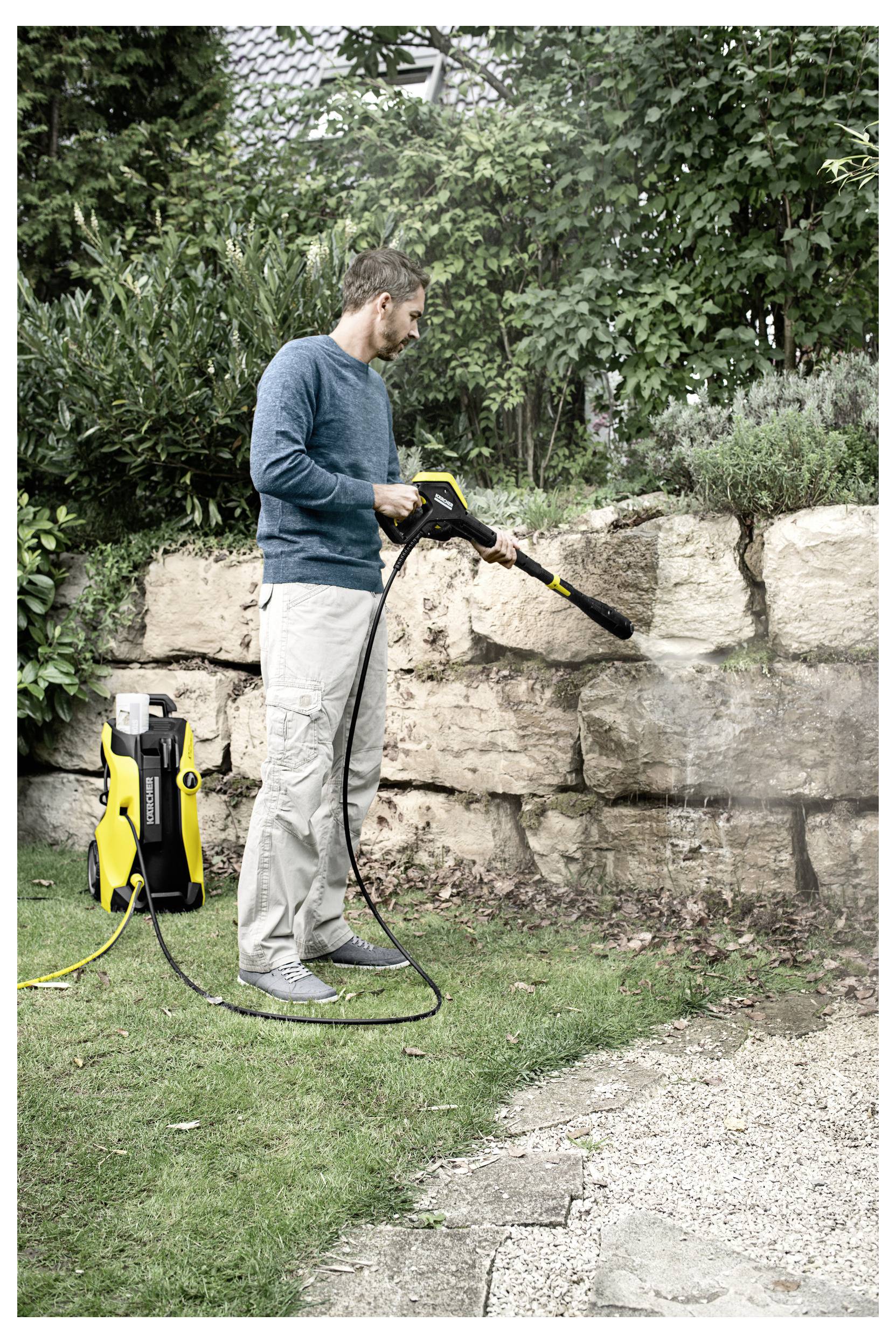 A person uses a pressure washer to clean a stone wall in a garden, surrounded by greenery and a house visible in the background.