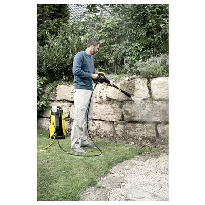 A person uses a pressure washer to clean a stone wall in a garden, surrounded by greenery and a house visible in the background.
