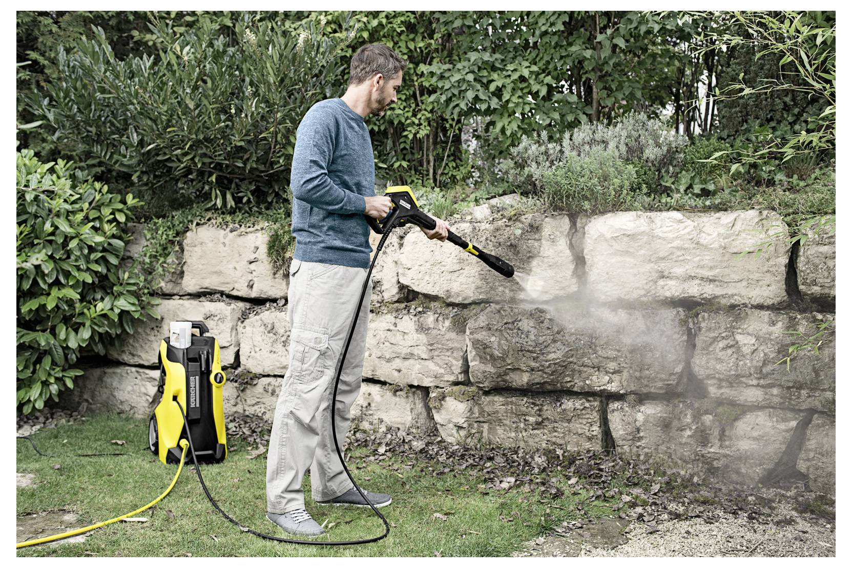 A person uses a pressure washer to clean a stone wall in a garden, surrounded by greenery and bushes.