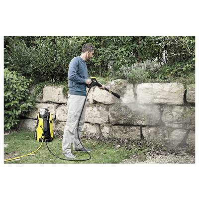 A person uses a pressure washer to clean a stone wall in a garden, surrounded by greenery and bushes.