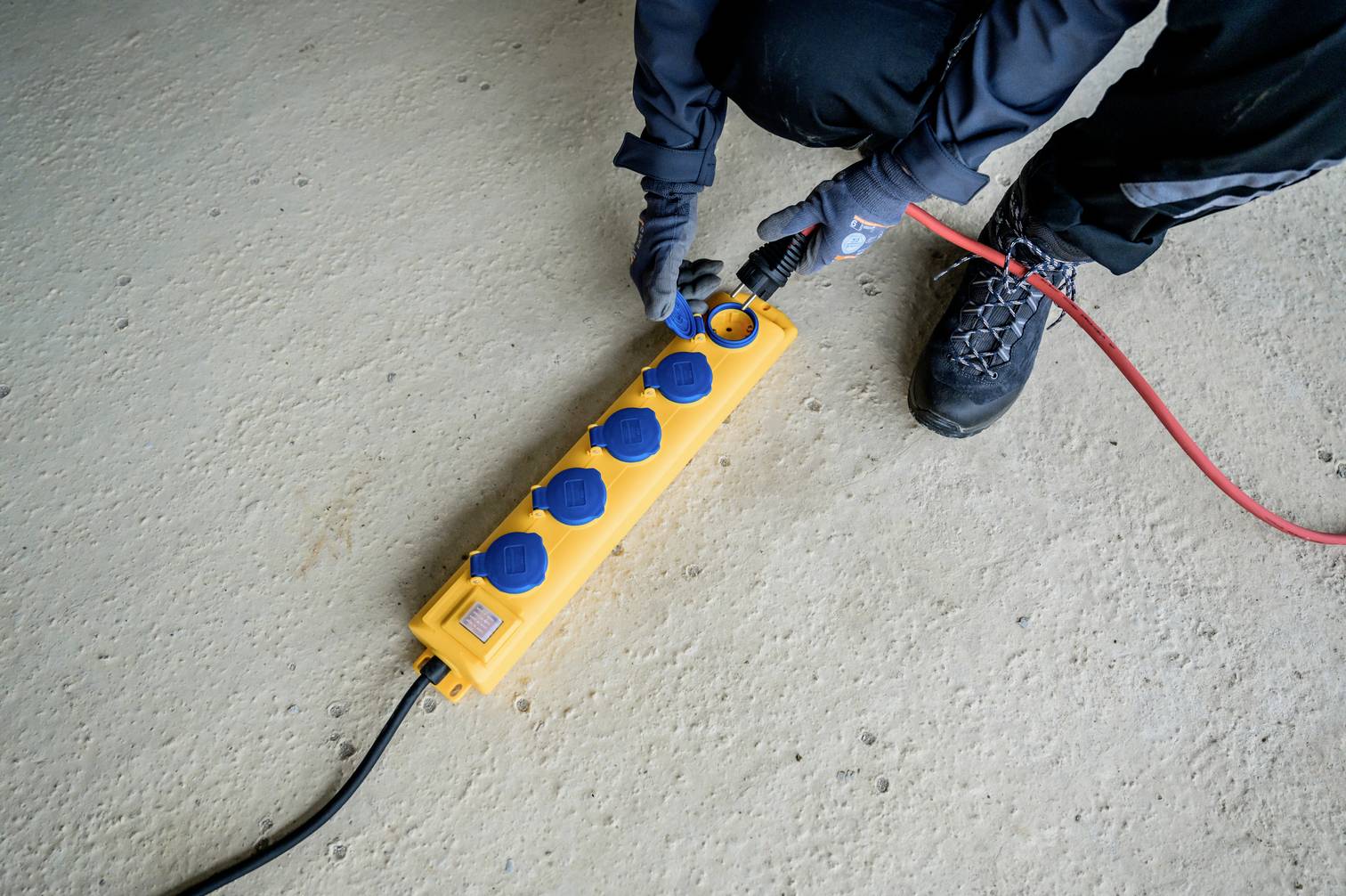 A person is plugging a red cable into a yellow power strip lying on a concrete floor. They are wearing gloves and work clothing.