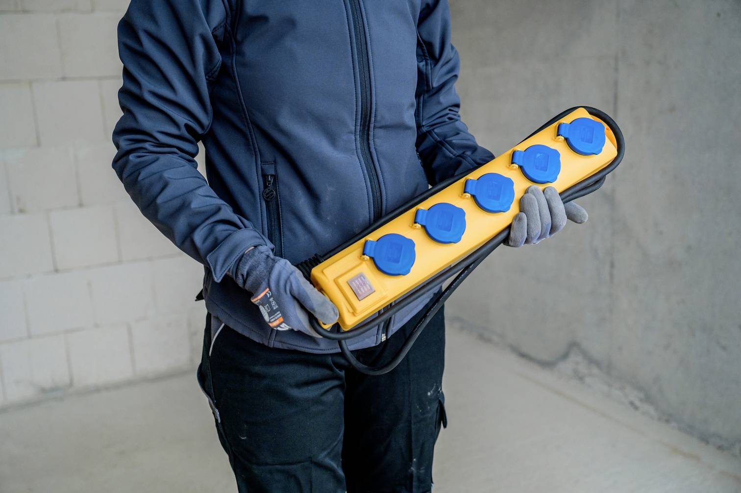 A person is holding a yellow power strip with five blue sockets. They are wearing a dark jacket and gloves. The background is an unfinished room.