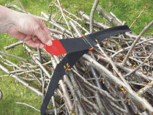A hand holds a tie-down strap, binding together a stack of cut branches. The branches are lying on a meadow.