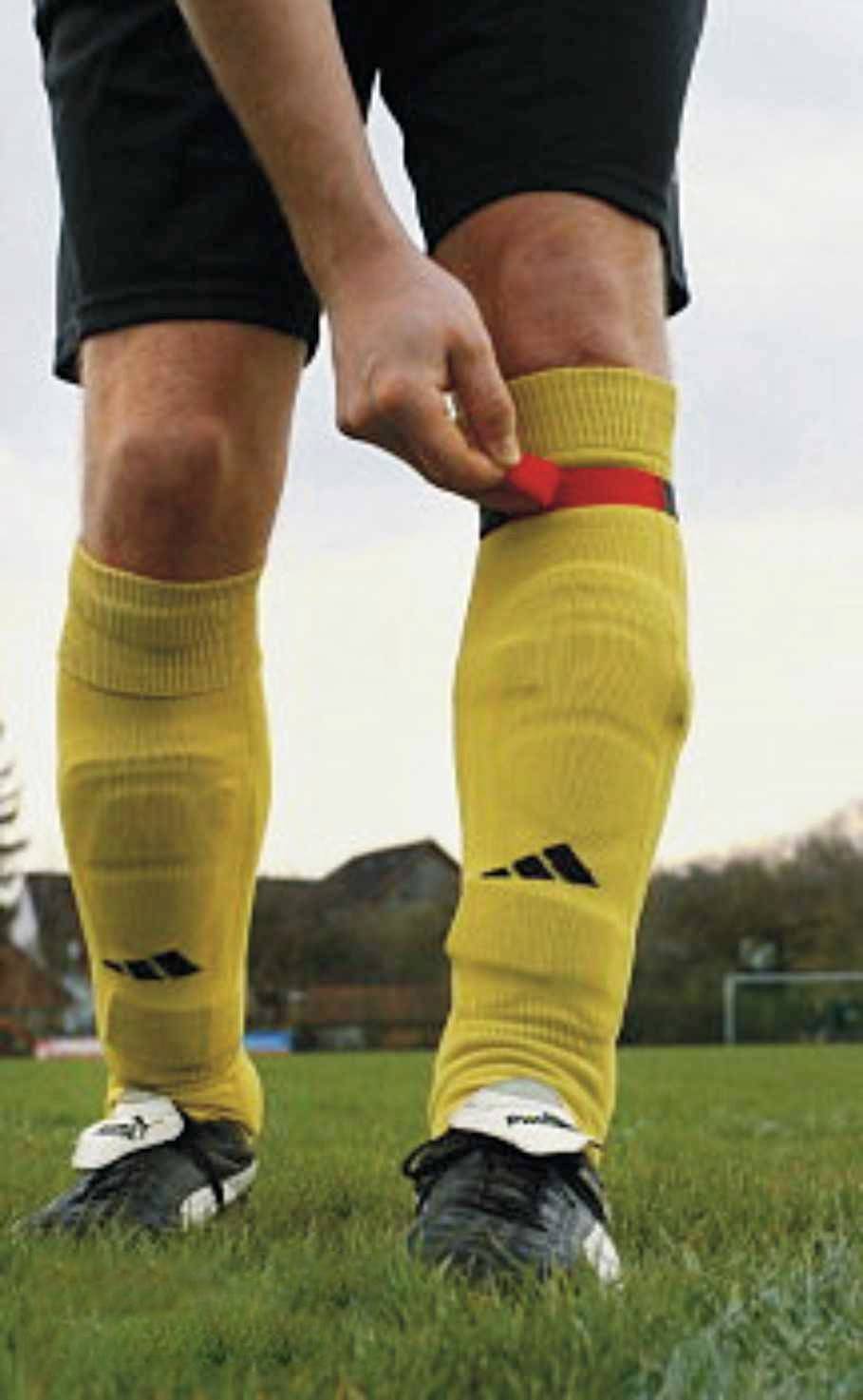 A player wearing yellow socks tightens a red sock tape over their ankle on a football pitch.