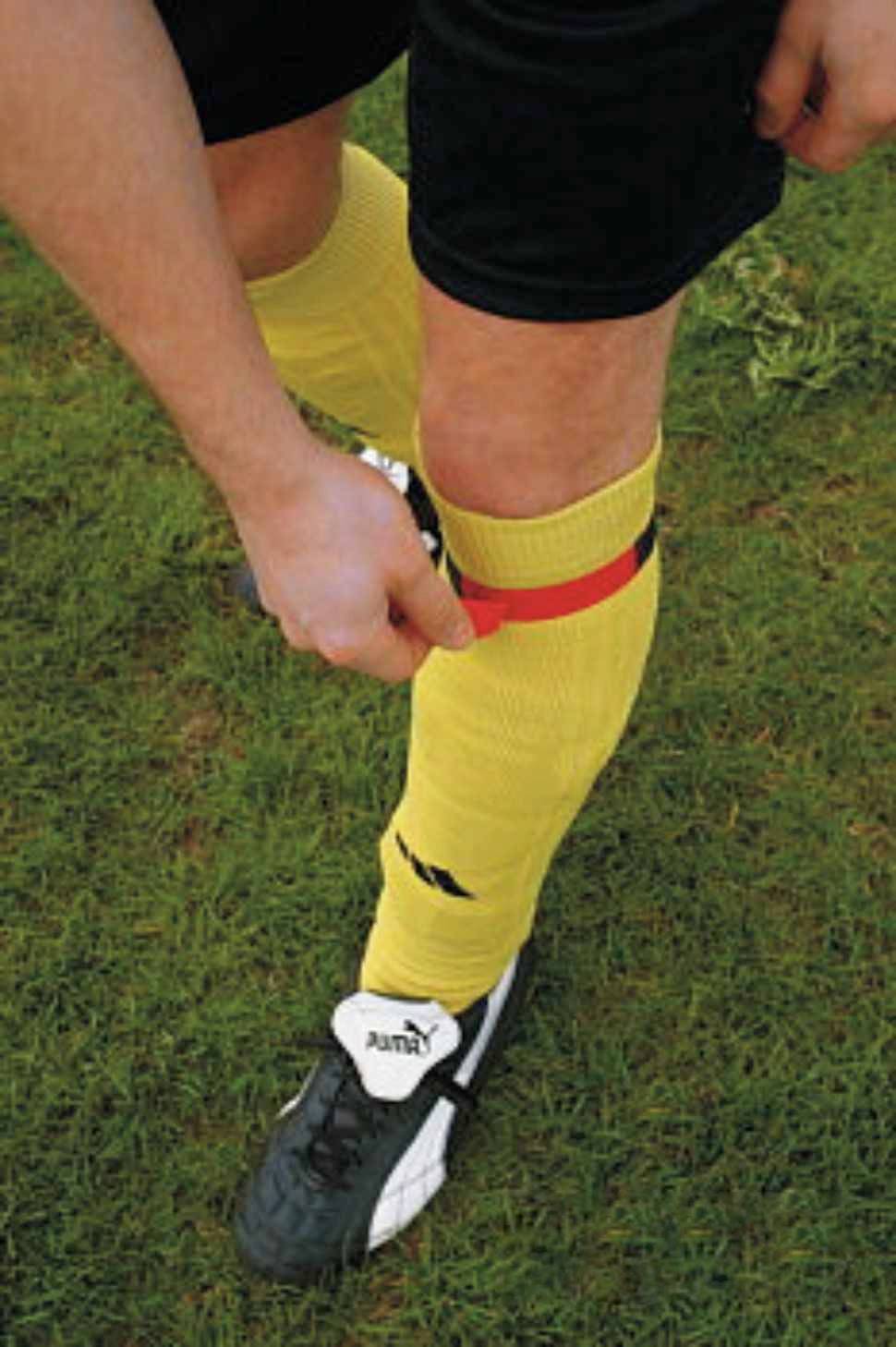 A person is wearing yellow socks and black football boots on a grass pitch. The person is fastening a red band around their left leg.