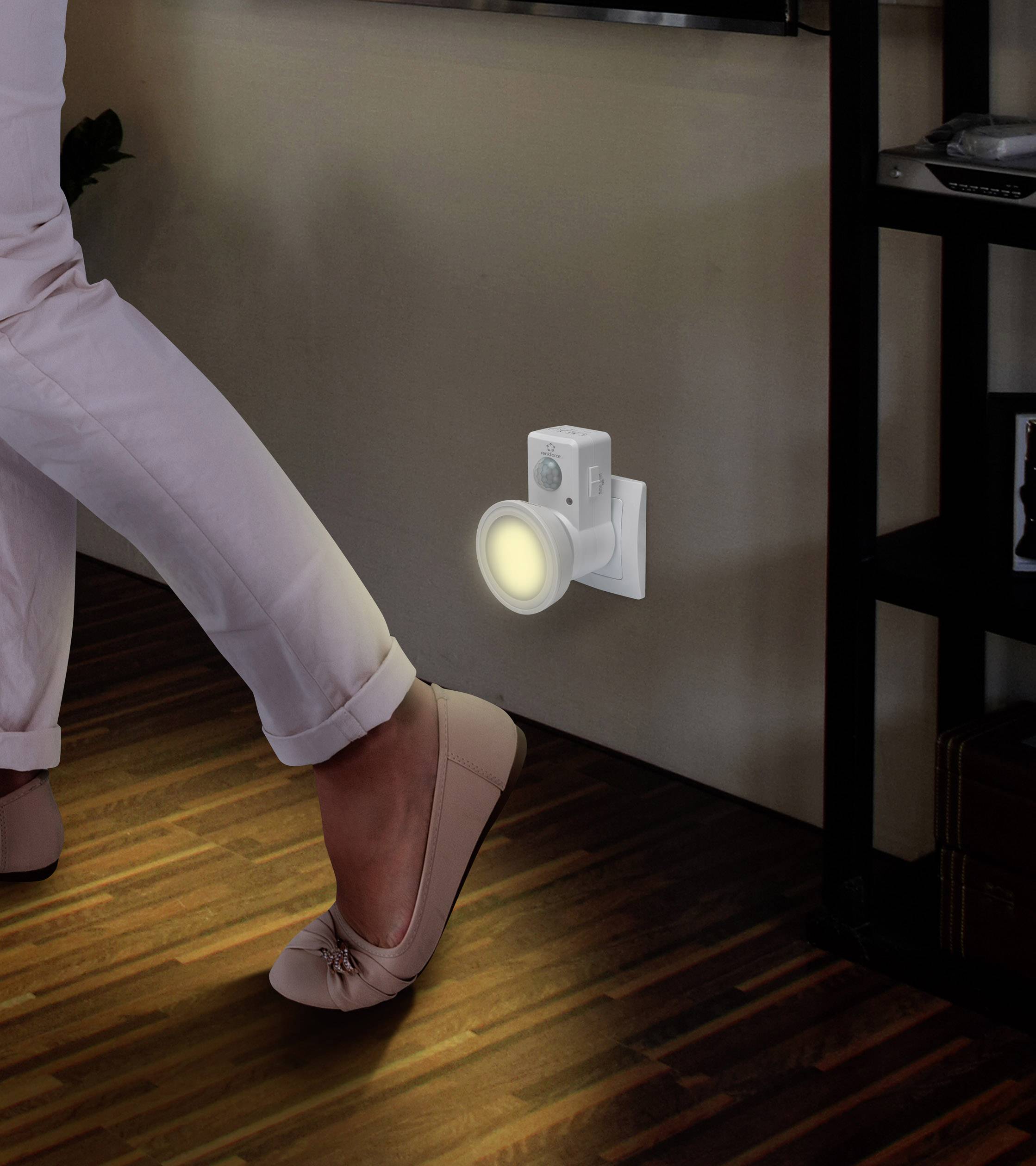 A person wearing white trousers walks past a plug-in night light stuck in the wall. The light illuminates the floor.