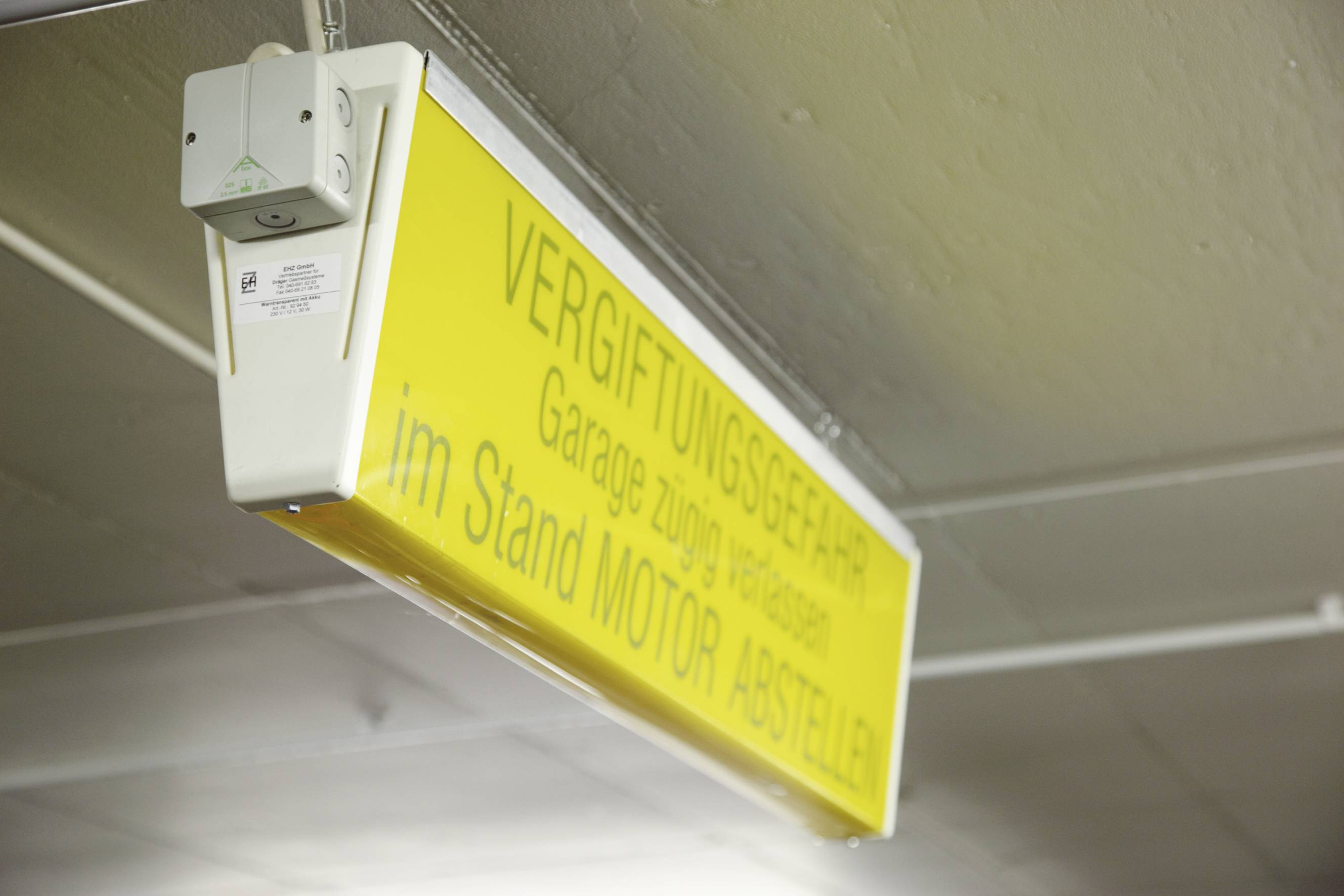 Yellow warning sign in a garage: 'Poison hazard. Ventilate garage additionally. Switch off engine when stationary.' Affixed to the ceiling.
