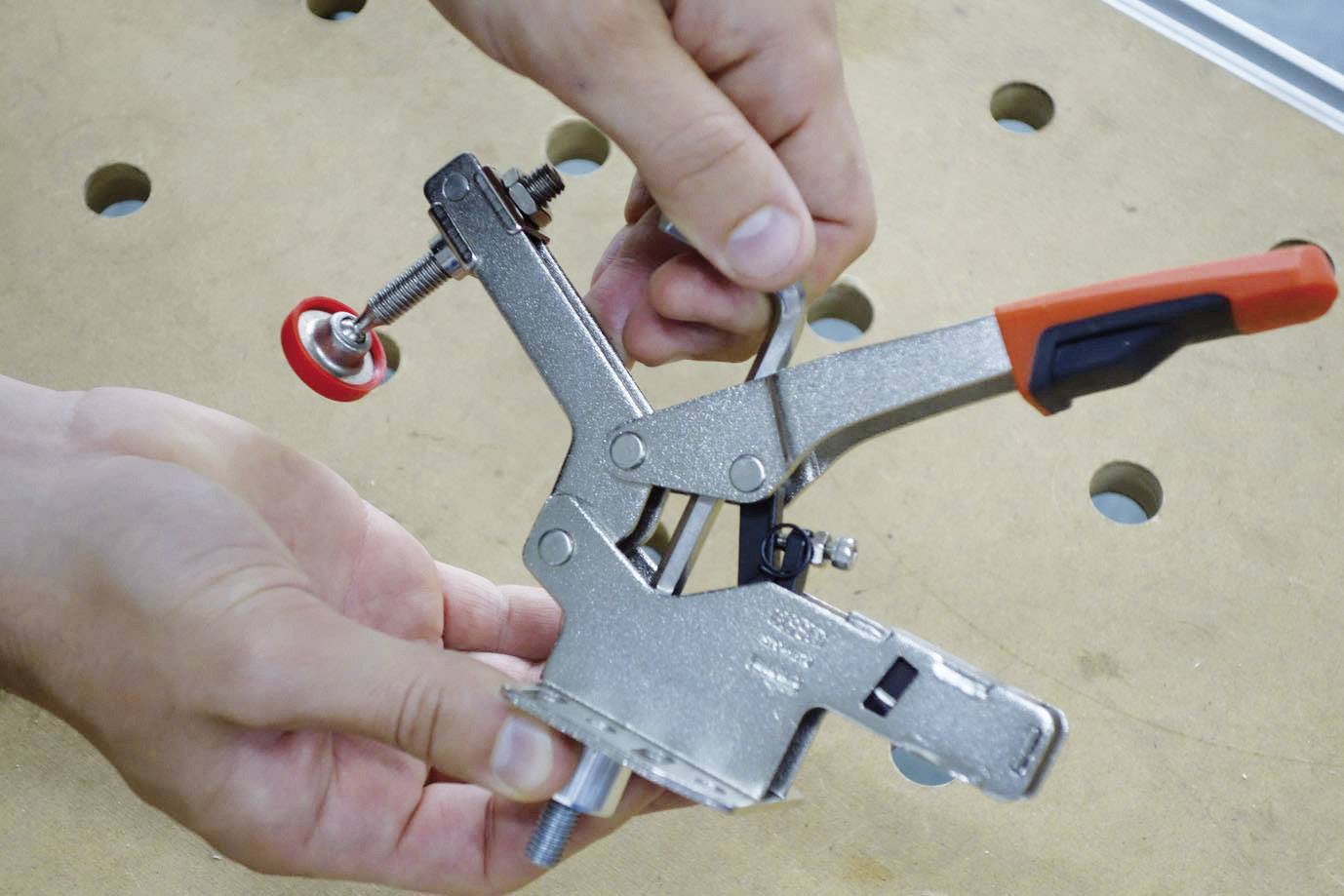 A person is holding a metal clip with red and black handles over a wooden table with holes.