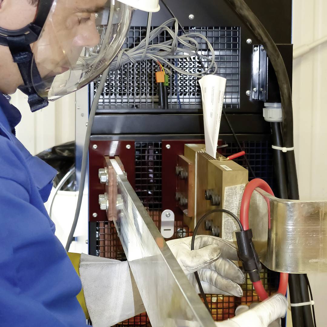 A worker is wearing protective clothing and working with a large electrical device. Technical machinery and cables are visible in the background.