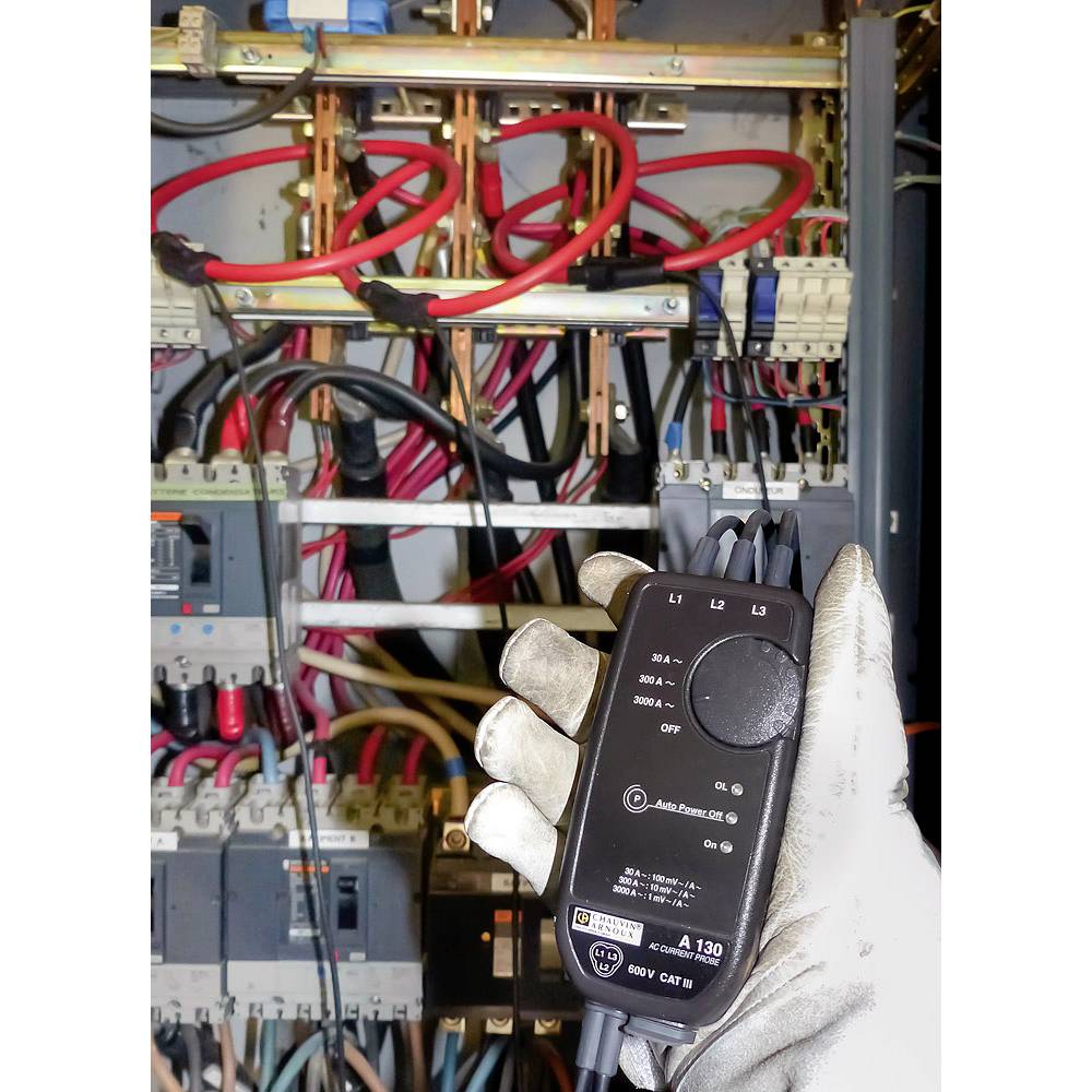 A gloved hand holds an electrical testing device in front of an open electrical cabinet with various cables and fuses.