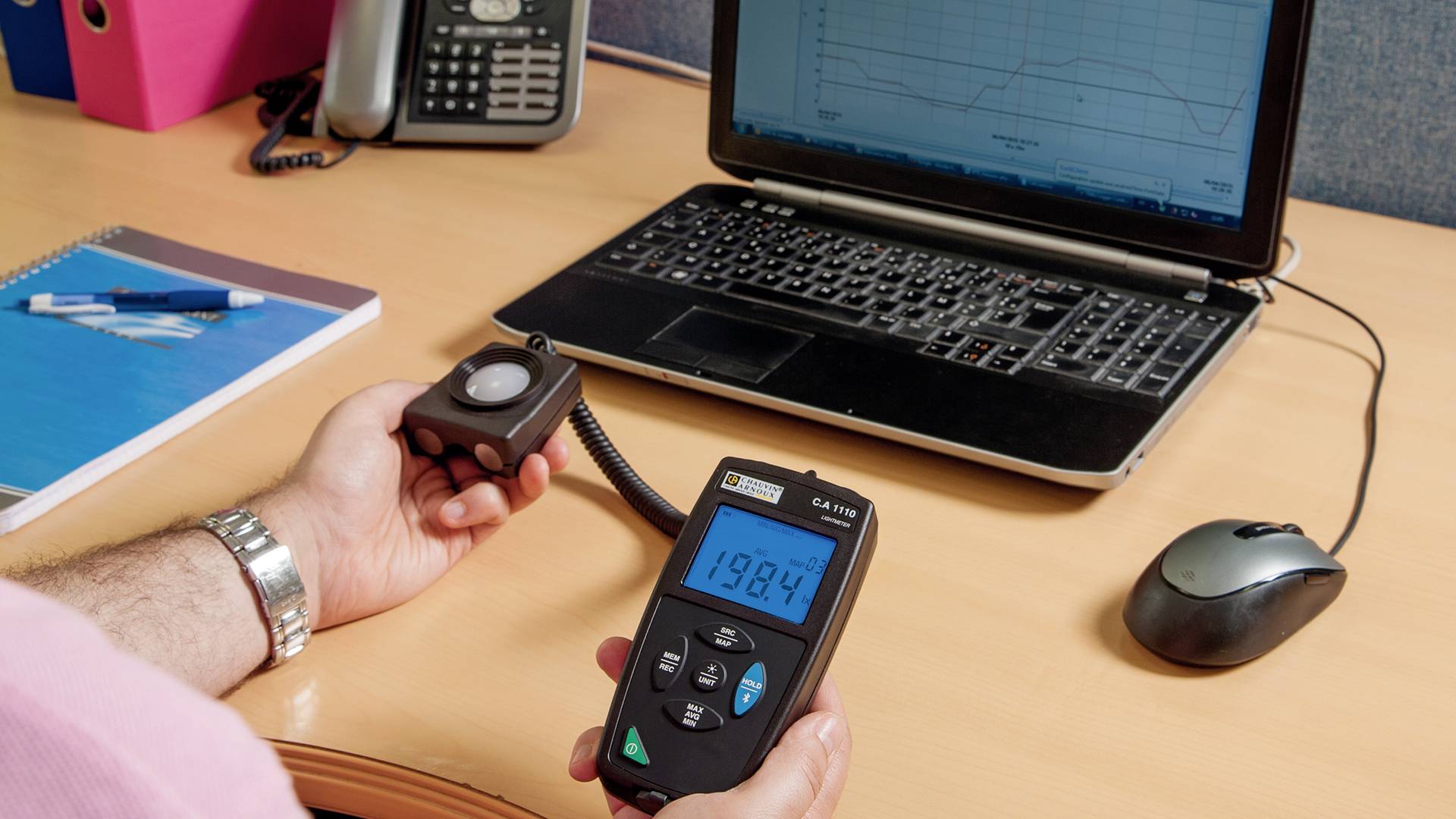 A person is holding a light meter displaying 198.4 in front of a laptop with a diagram on the screen in an office.