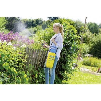 A woman is watering plants in a garden with a yellow and blue sprayer. She is standing next to a wooden fence with flowering shrubs.