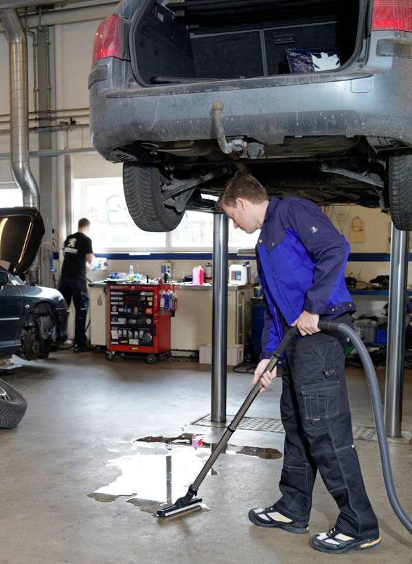 A mechanic uses a vacuum to clean a spill on the workshop floor under a car on a lift. Another mechanic works at a tool bench in the background.