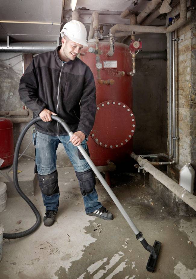 A person in a construction site wearing a hard hat and protective clothing uses an industrial vacuum to clean debris from the floor.