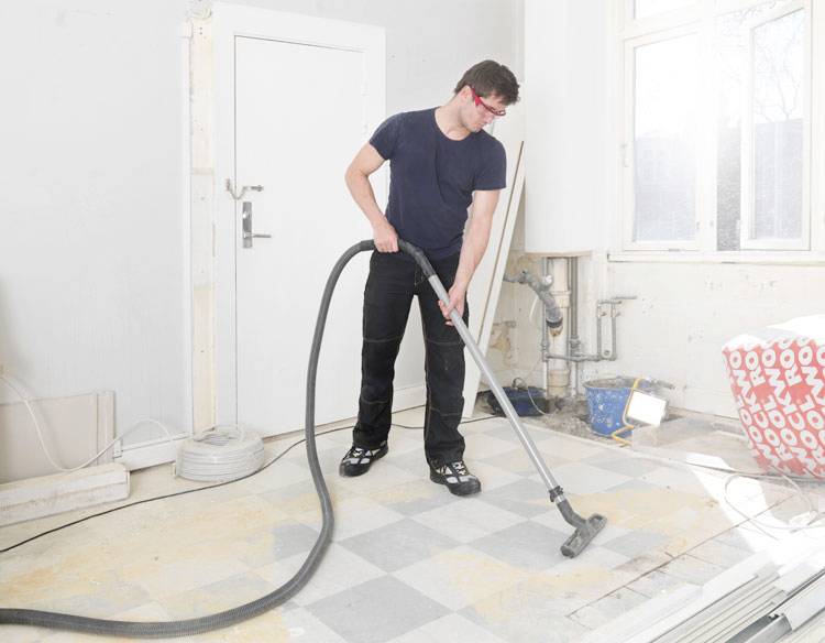 A person in a construction setting vacuuming a dusty floor. The room is under renovation, with tools and construction materials visible.